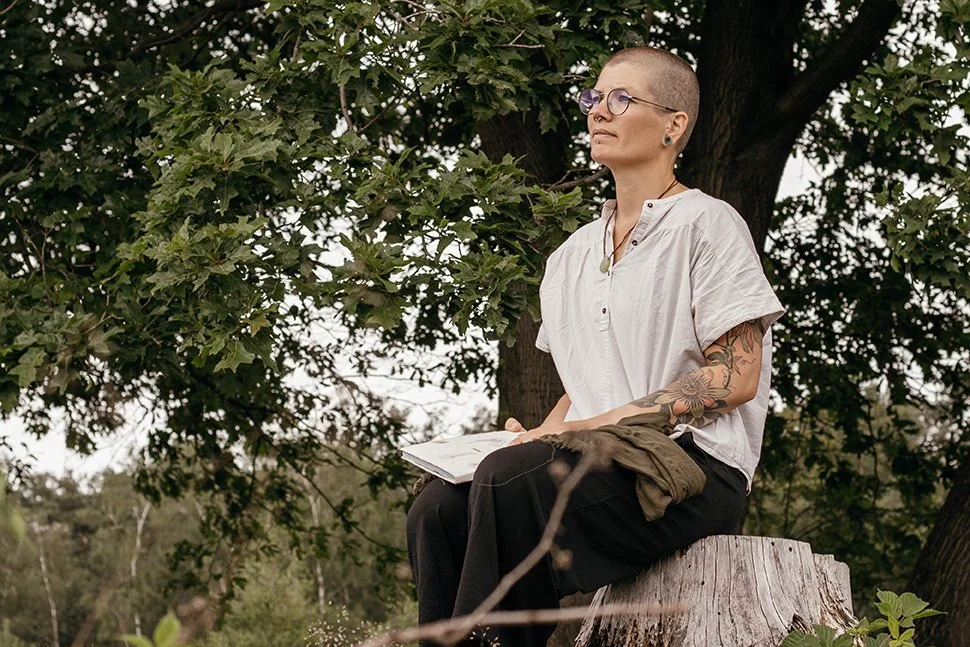 A woman with short hair, glasses, and tattoos, sitting on a tree stump outdoors, holding an open book, with a tree and green foliage behind her.