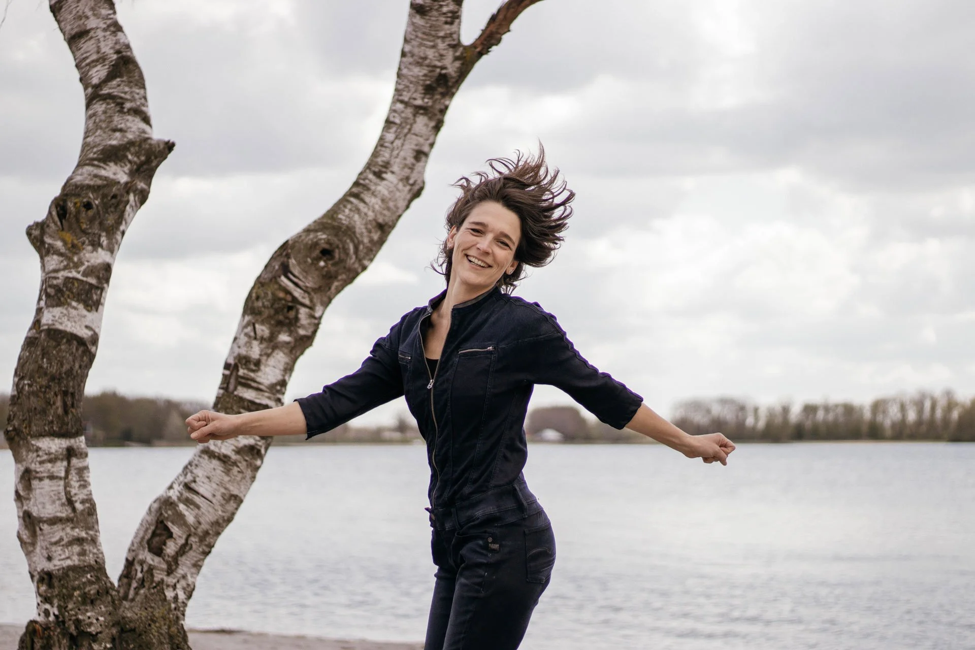 Woman smiling and jumping near a tree by a lake on a cloudy day.