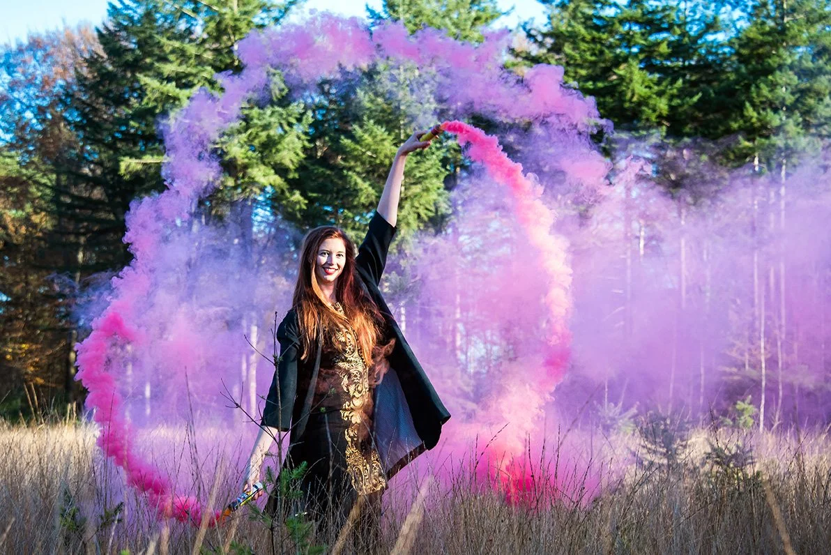 A woman holding a pink smoke grenade, creating a rainbow-shaped smoke ring in a field with tall grass and trees in the background.