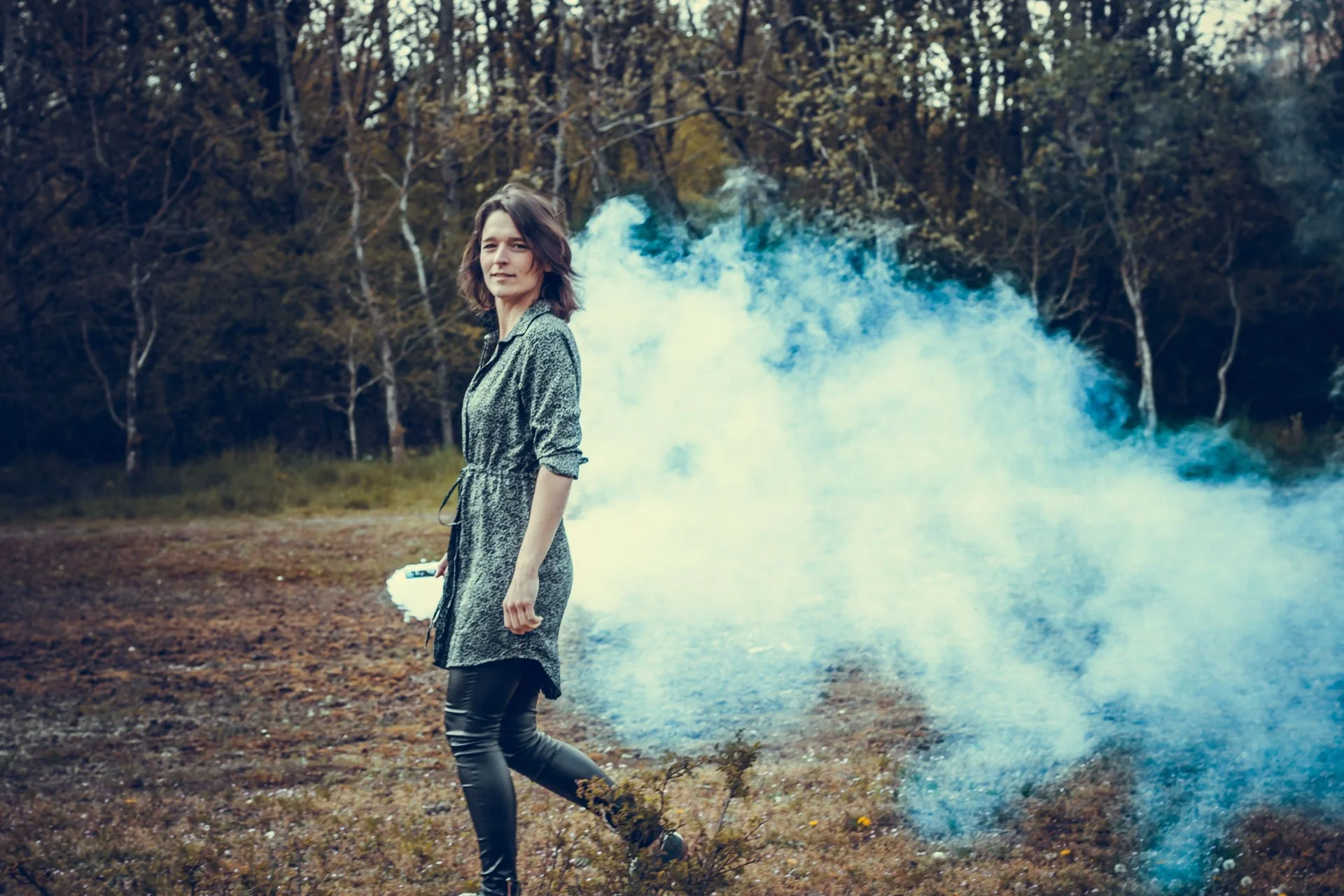 Woman standing in a forest at dusk with smoke behind her.