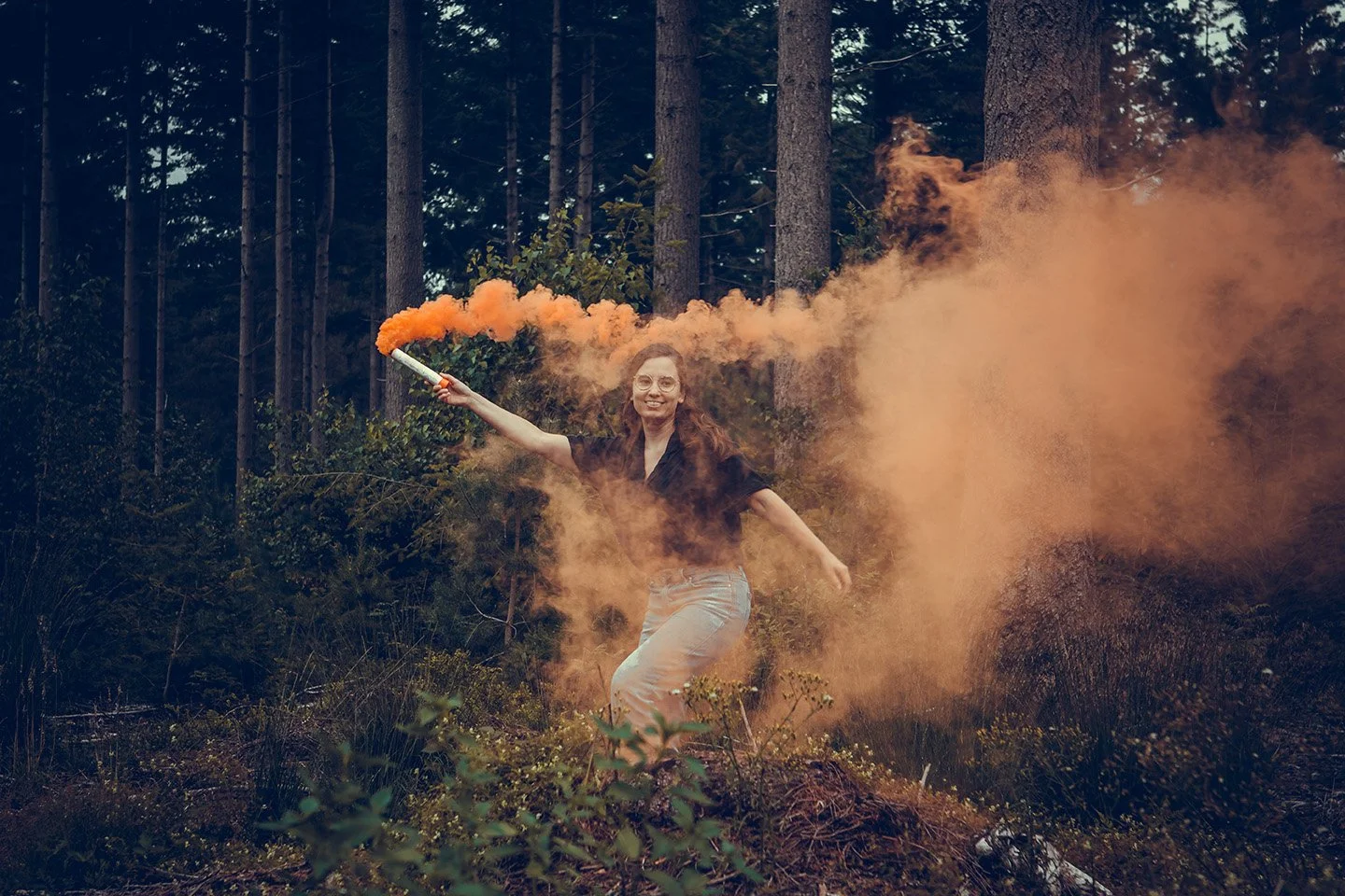 A woman smiling and holding an orange smoke flare in a forested area, with orange smoke billowing behind her.