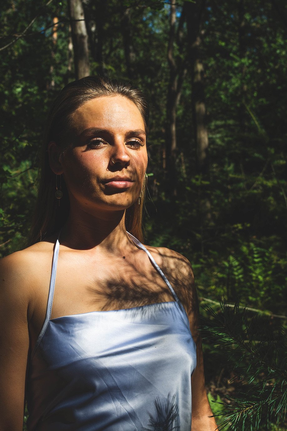 A woman standing in a sunlit forest, wearing a light-colored, sleeveless top, with shadows of leaves on her face and shoulder.