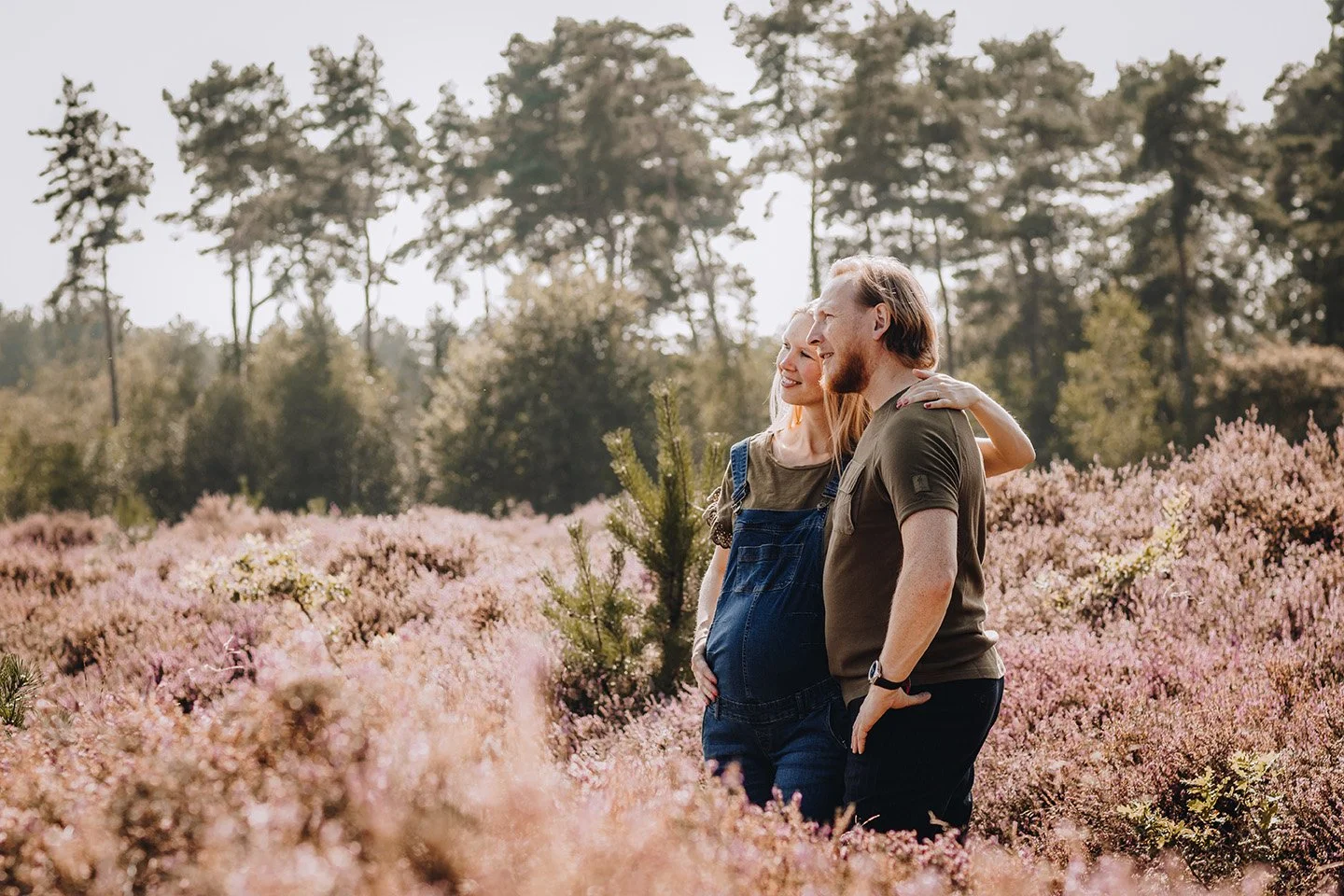 A pregnant woman and a man stand close together in a field of pink flowers, smiling and looking into the distance, with trees in the background.