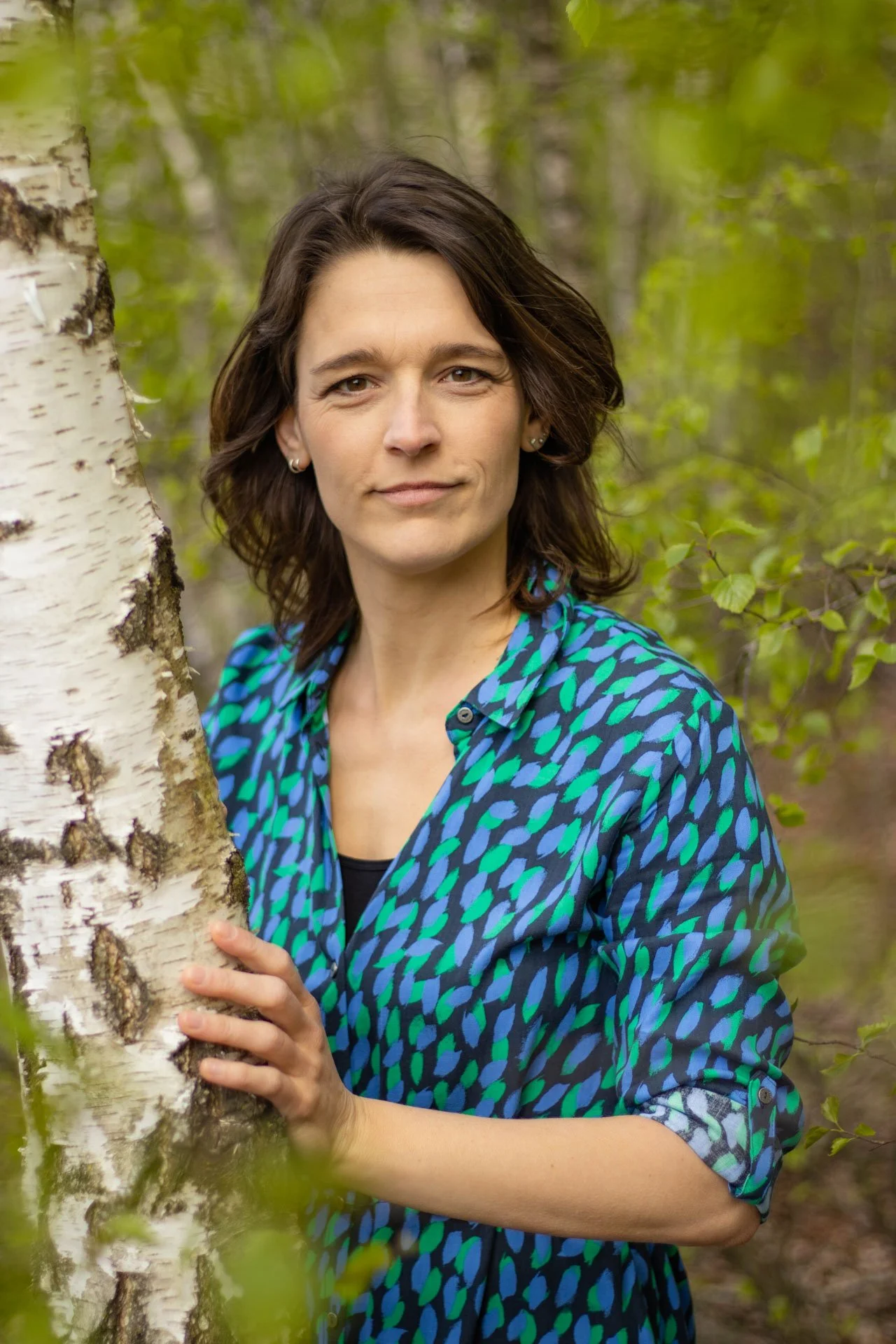 A woman with dark brown hair and light skin holding a white birch tree in a green forest.