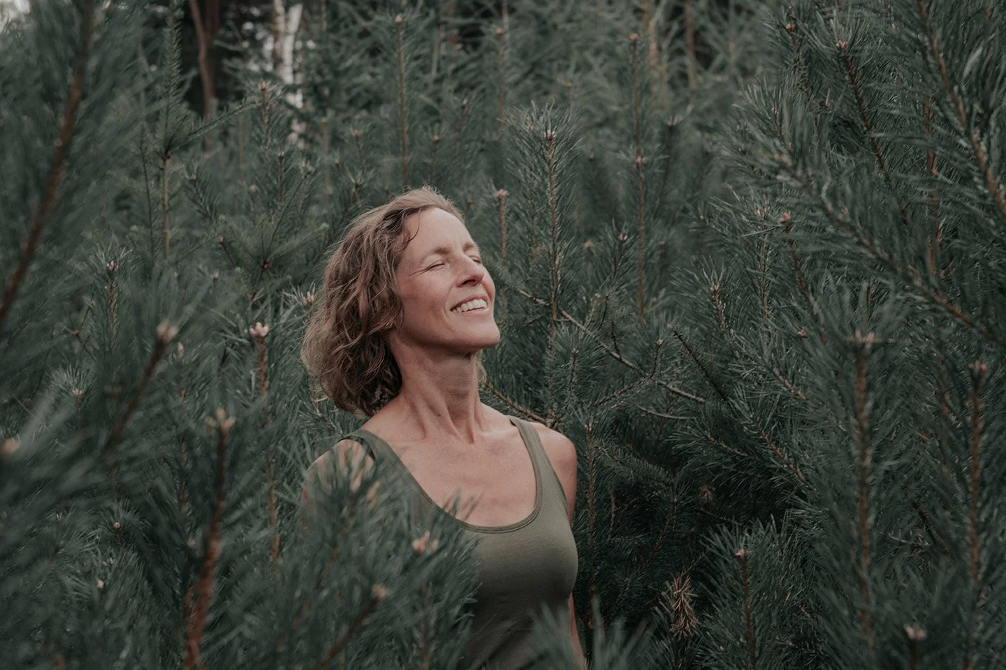 A woman in a green tank top standing among green pine trees with her eyes closed and a joyful smile.