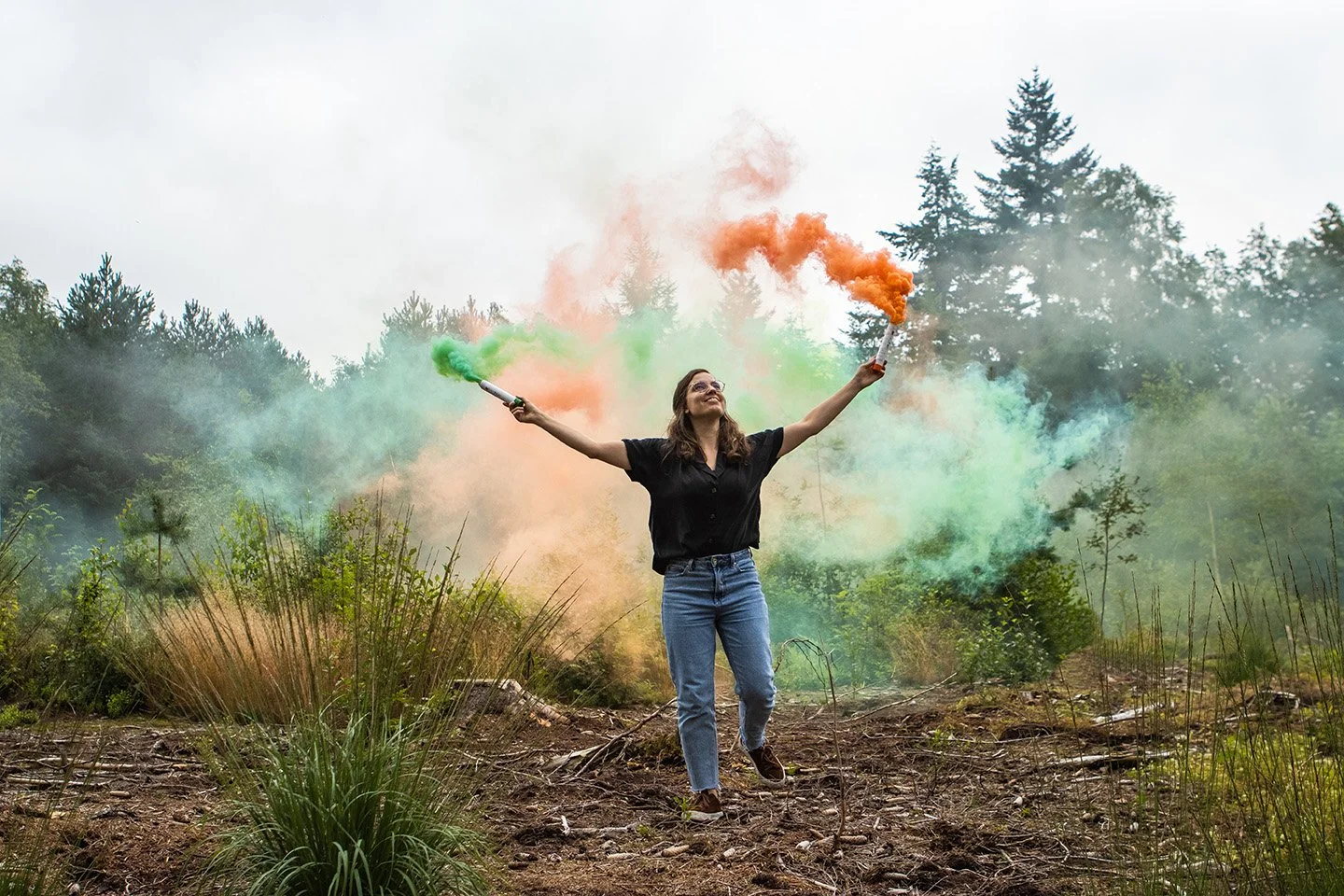 Woman smiling and running in a forest clearing, holding two smoke flares emitting orange and green smoke, with trees in the background.