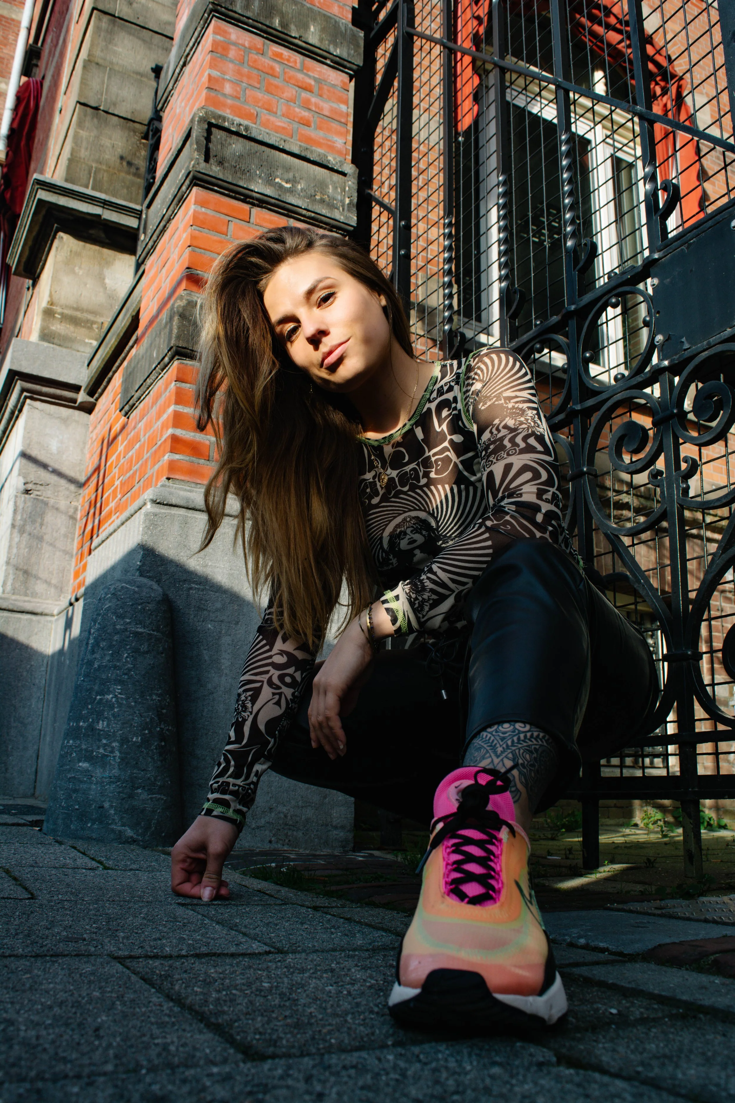 A young woman with long brown hair squatting on a city sidewalk, pointing her finger at the ground, with her face angled toward the camera, wearing a patterned long-sleeve shirt, black leather pants, and pink and yellow sneakers, next to a black wrought-iron gate and a brick building.