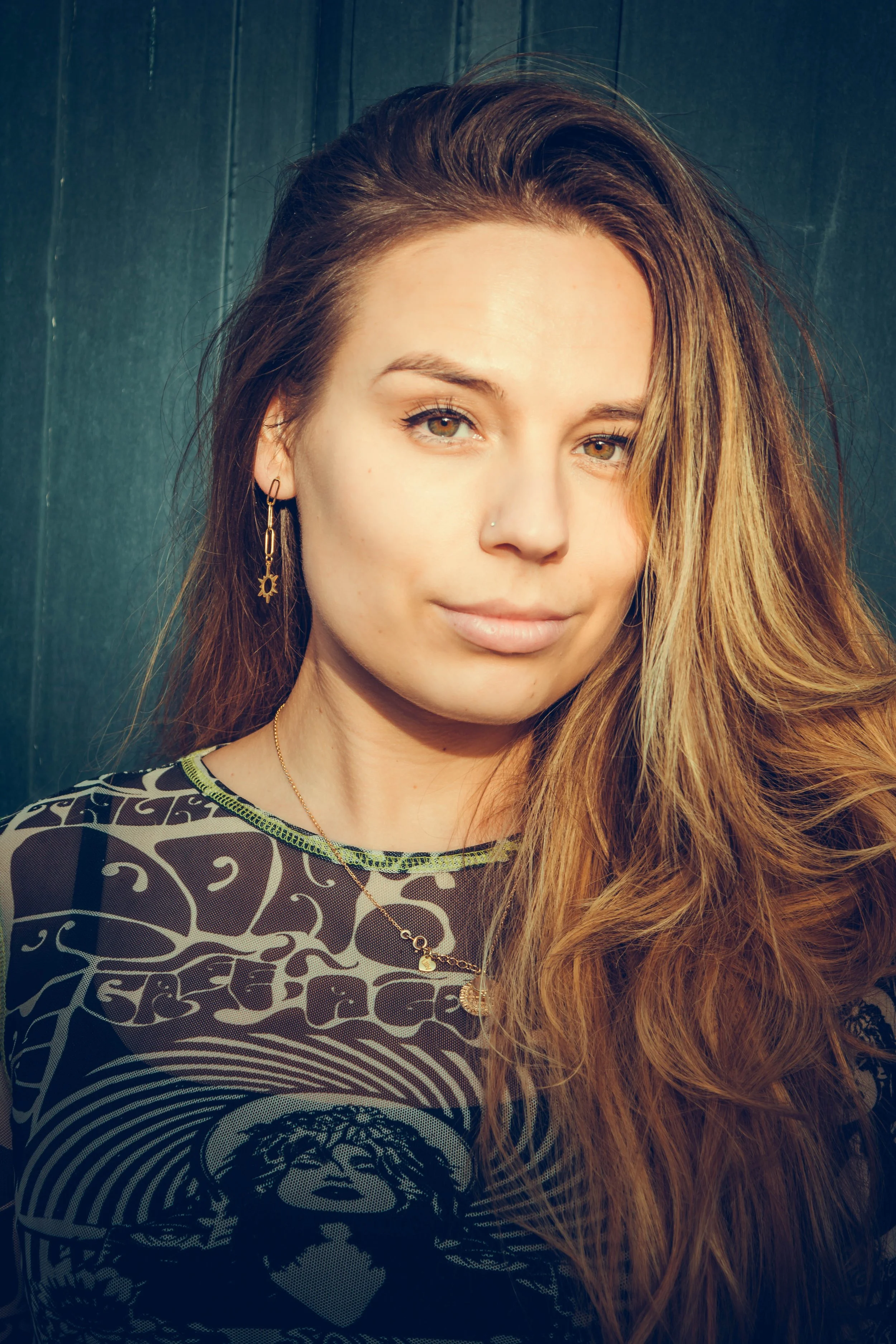 Close-up of a young woman with long, wavy blonde hair, wearing earrings and a patterned top with a graphic design, standing against a dark background.