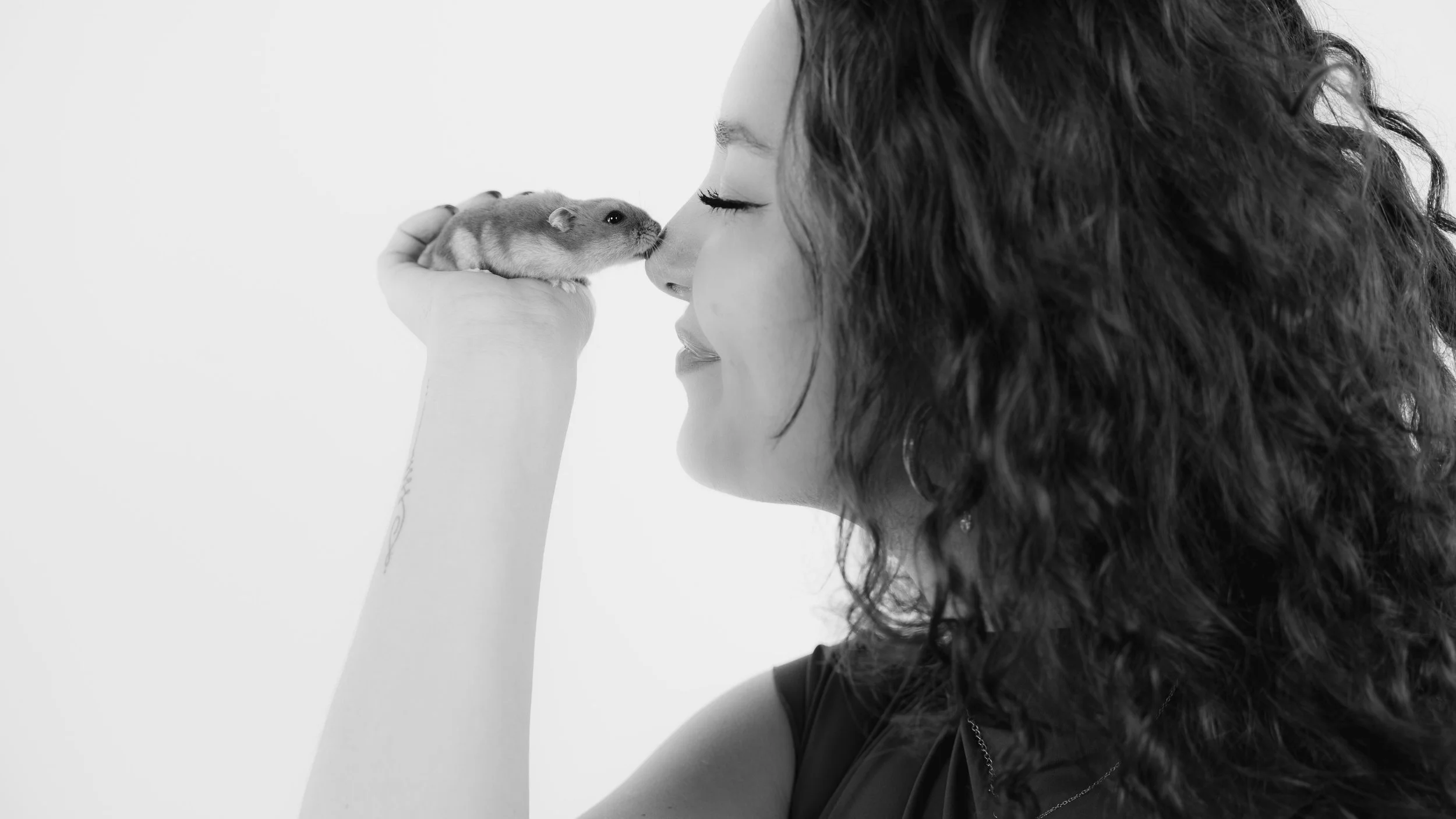 A woman with curly hair holding a small hamster close to her face, touching noses, in a black and white photo.