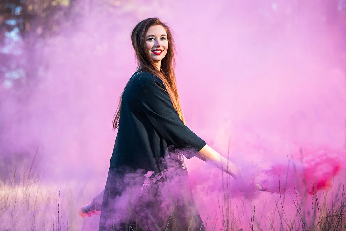 A woman with long brown hair smiling in a field of grass with pink and purple smoke.