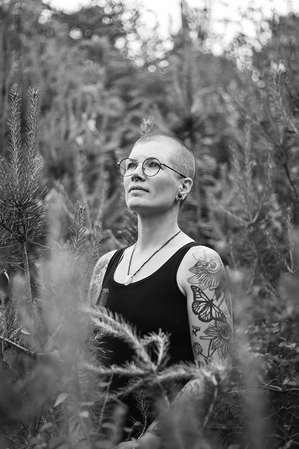 A woman with a shaved head and glasses stands among pine trees, looking upwards. She has tattoos on her right arm, wears a black tank top, glasses, and a necklace.