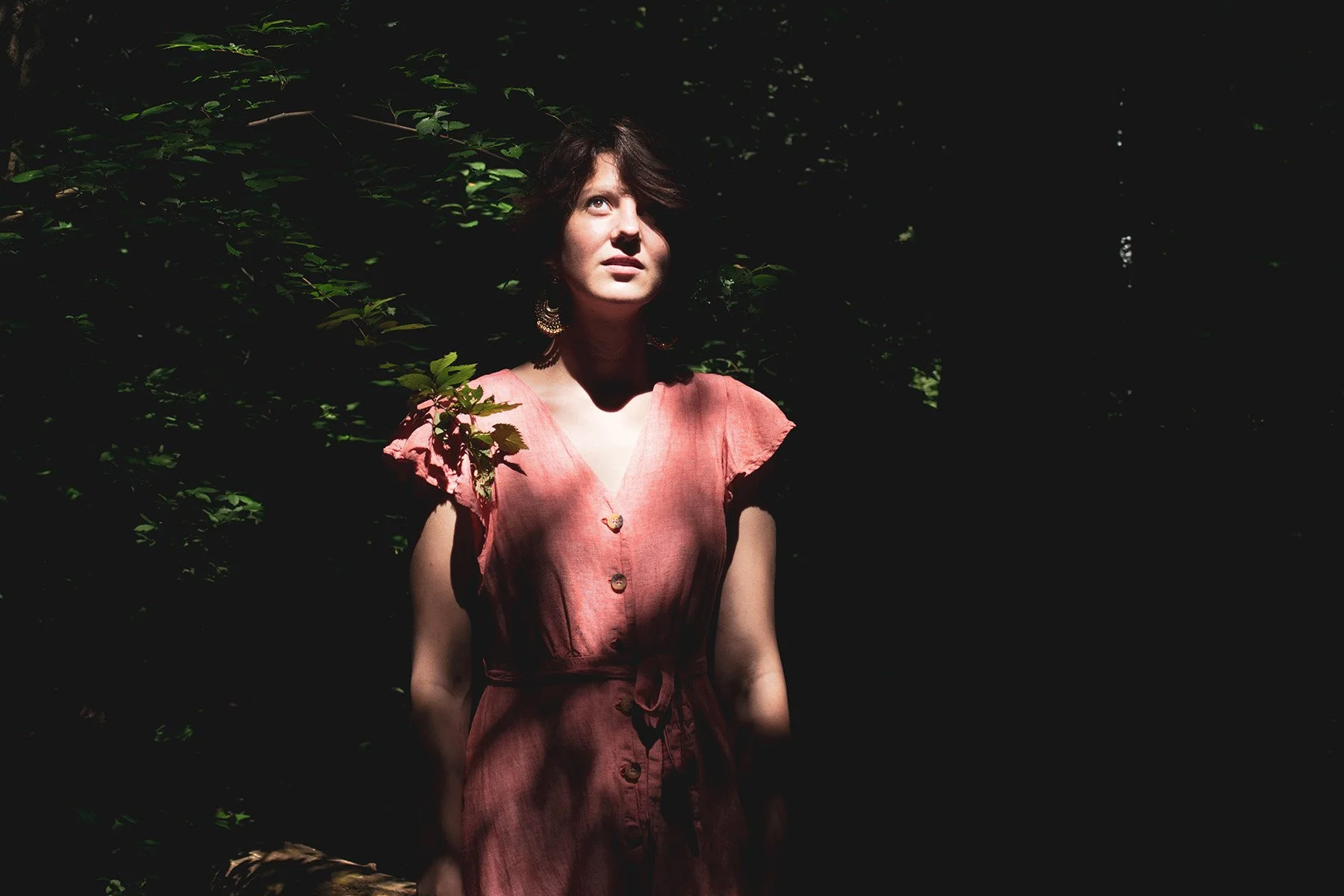 A woman with short dark hair wearing a pink dress standing in a forest with sunlight filtering through the leaves.