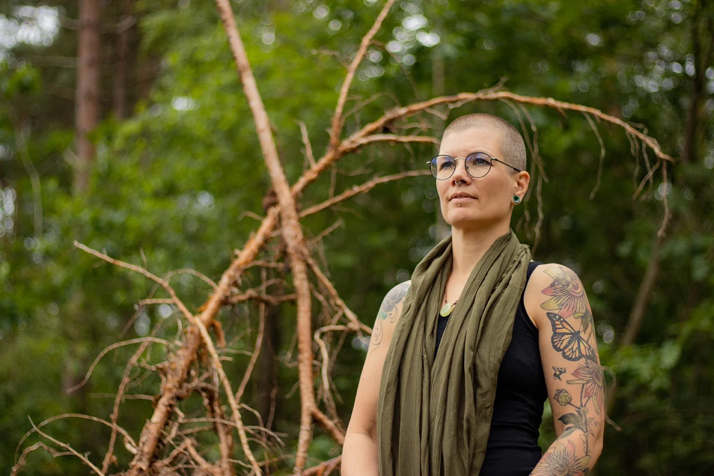 Woman with a shaved head, glasses, tattoos on arms, and wearing a green scarf around her neck, standing in a forested area with trees and branches.
