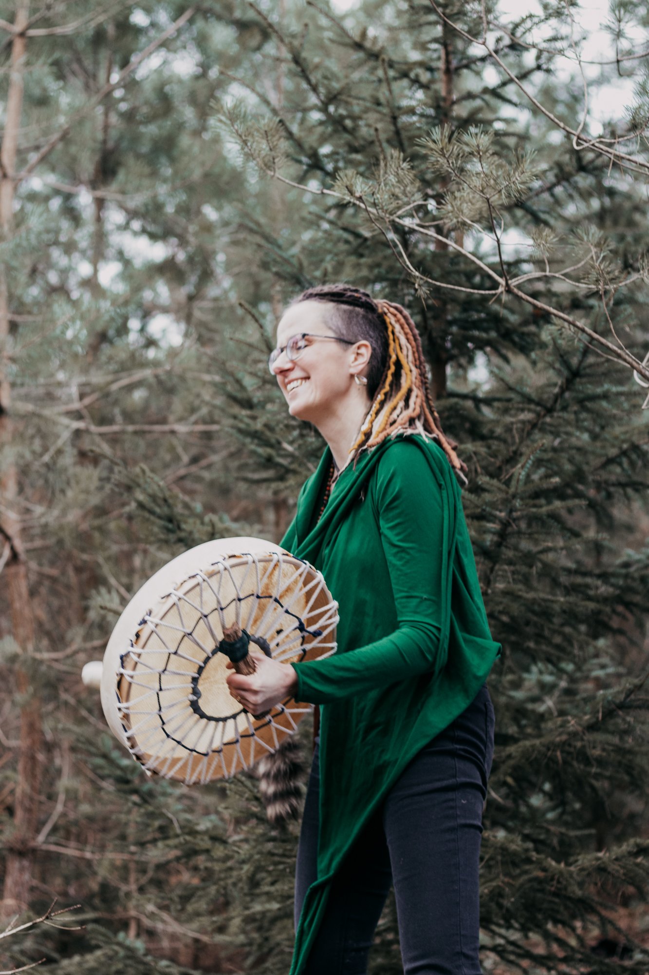 A woman with glasses and braided, colorful hair smiling in a forest while holding a drum