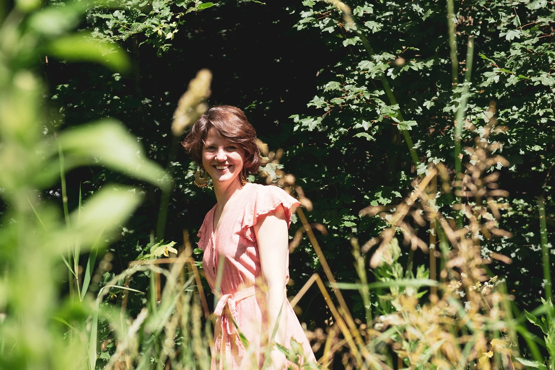 A woman smiling and wearing a pink dress in a lush green outdoor setting with tall plants and trees.