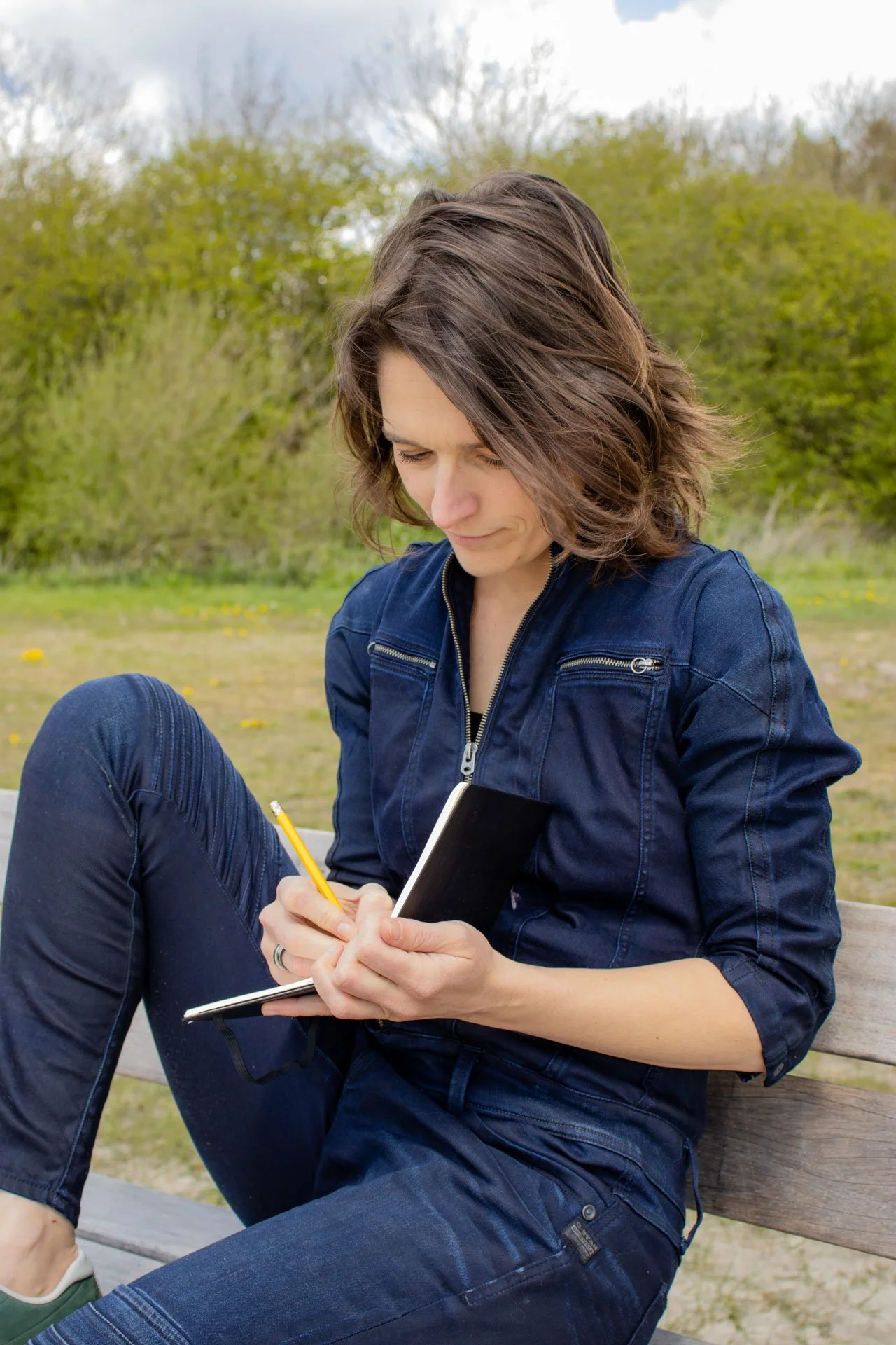 A woman sitting on a park bench, dressed in a denim jacket and jeans, holding a notebook and writing with a yellow pencil, with trees and a cloudy sky in the background.