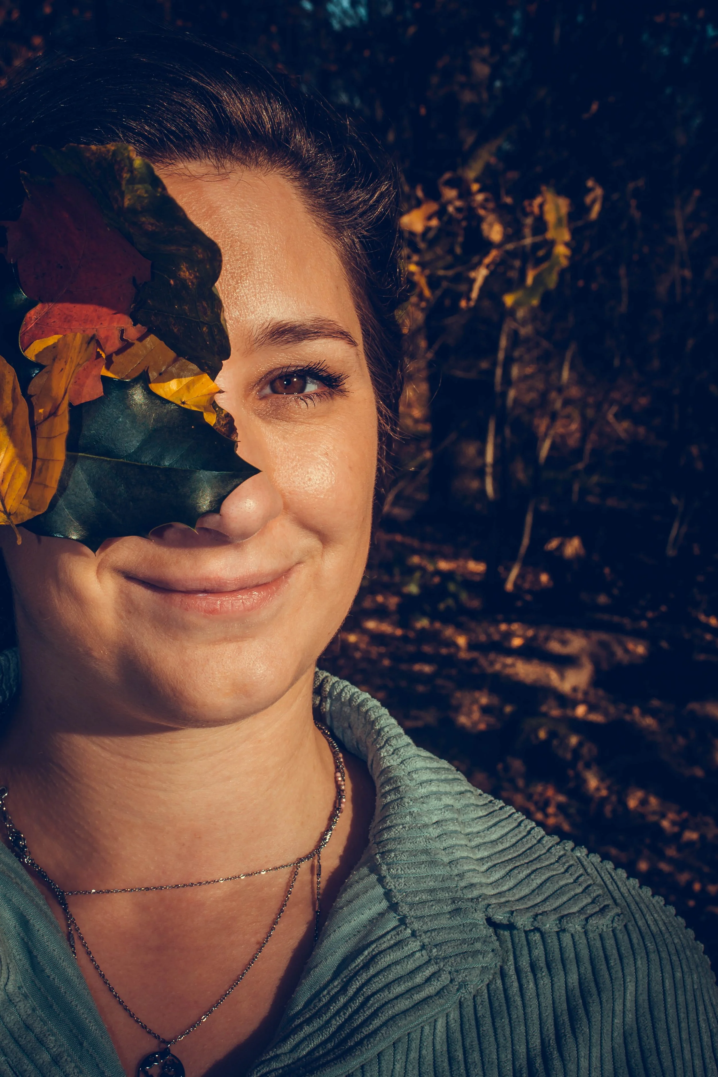 A woman smiling outdoors holding autumn leaves in front of her face, with one eye visible, in a wooded area during sunset.