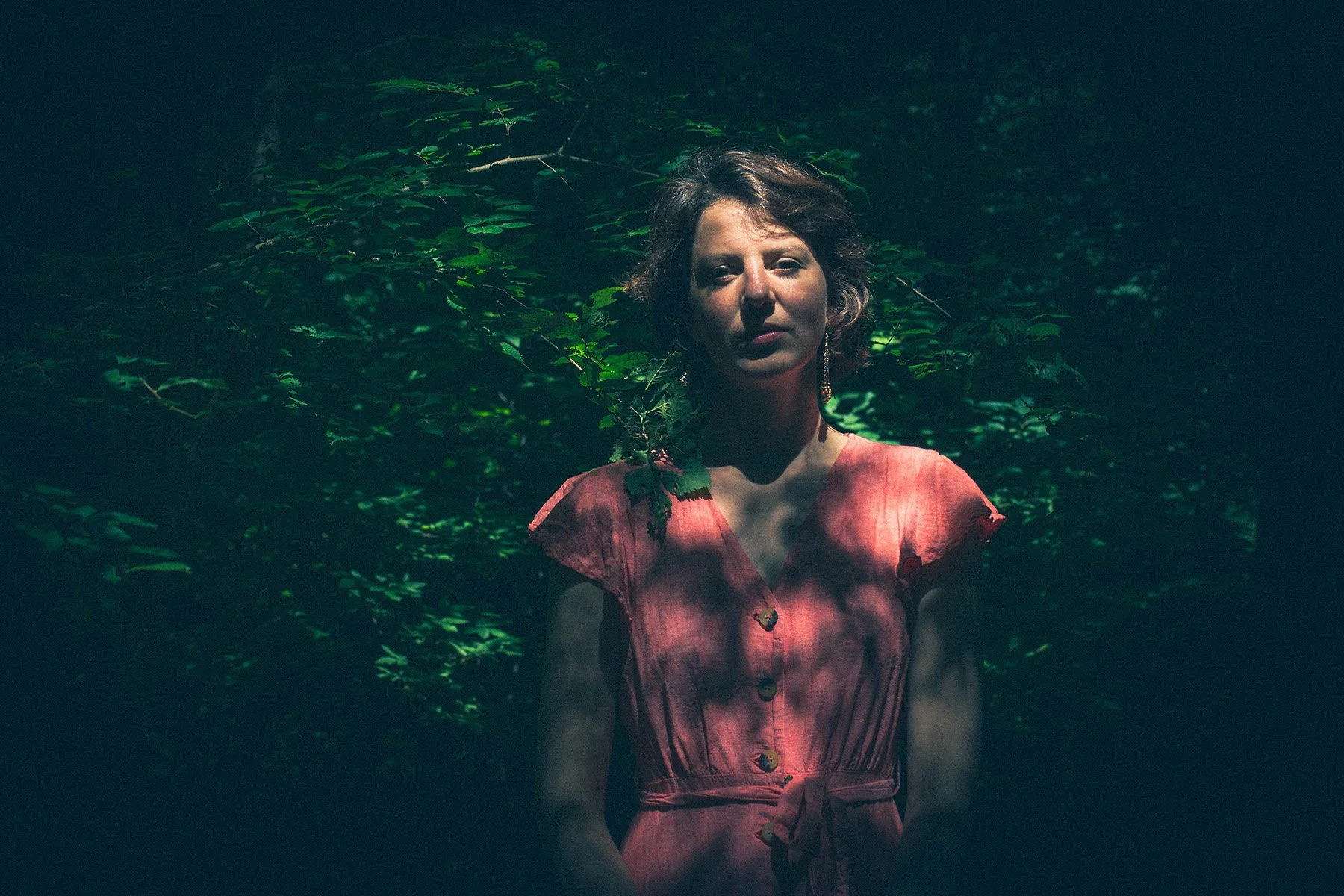 A woman in a pink dress stands in front of green foliage, illuminated by soft light with shadows across her face.