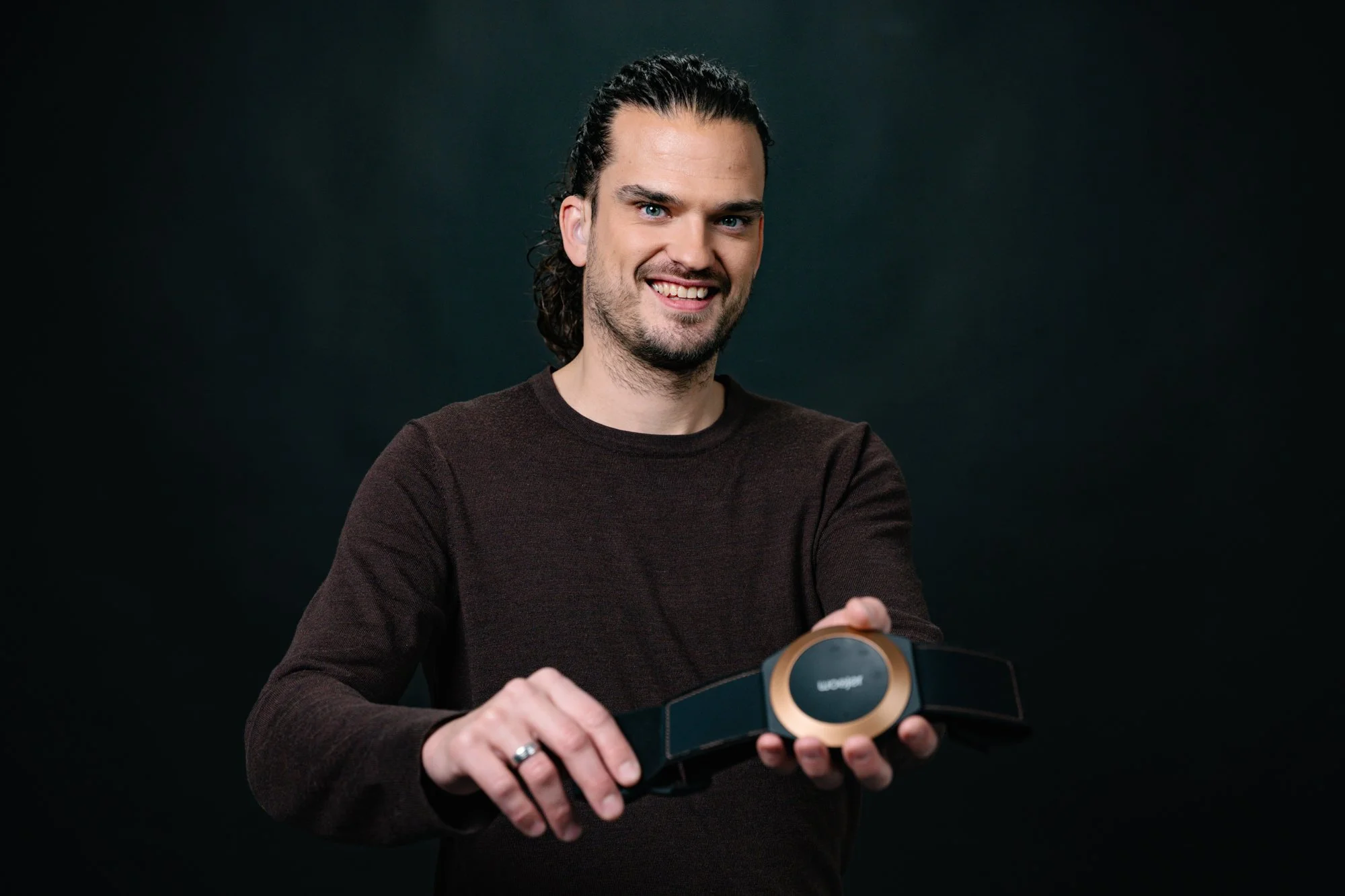 A man with long dark curly hair, wearing a dark brown sweater, is smiling and holding a black and gold virtual reality headset.