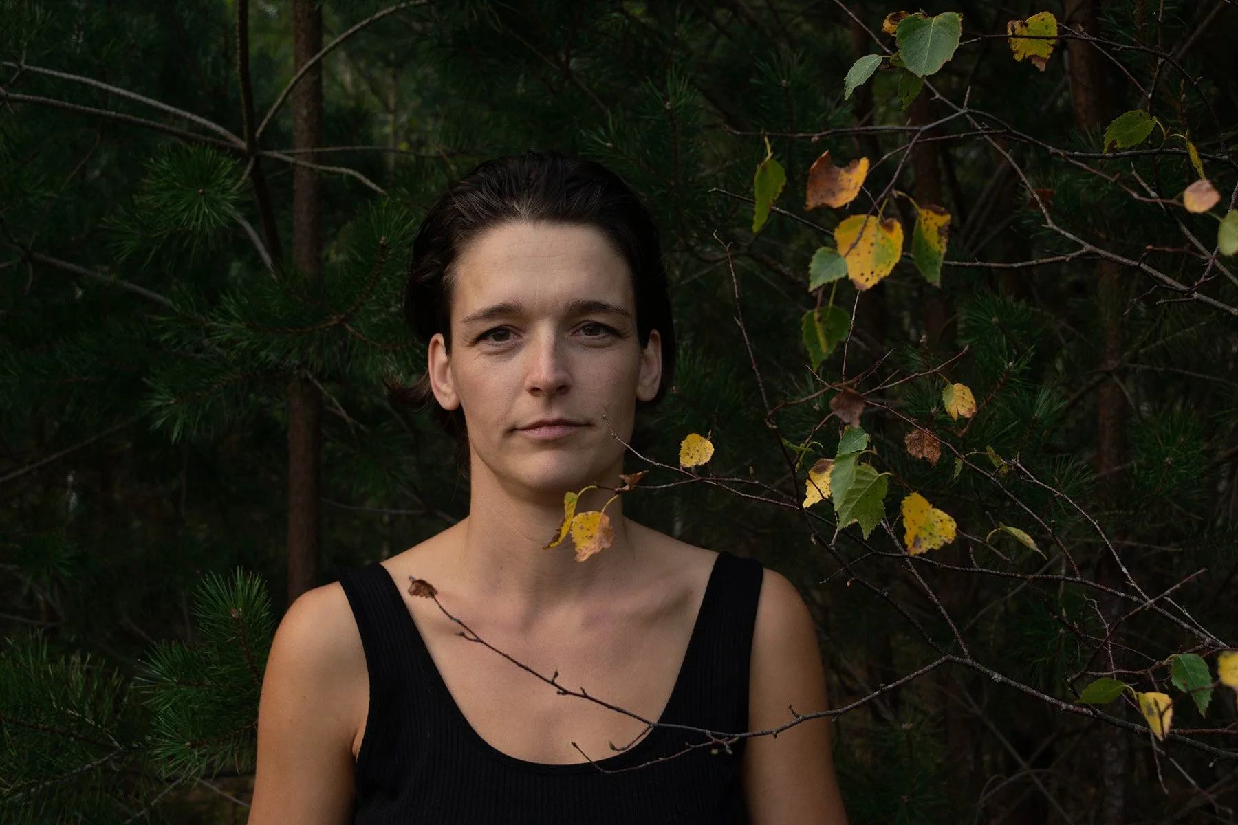 A woman with short dark hair and a black sleeveless top standing in front of greenery with leaves.