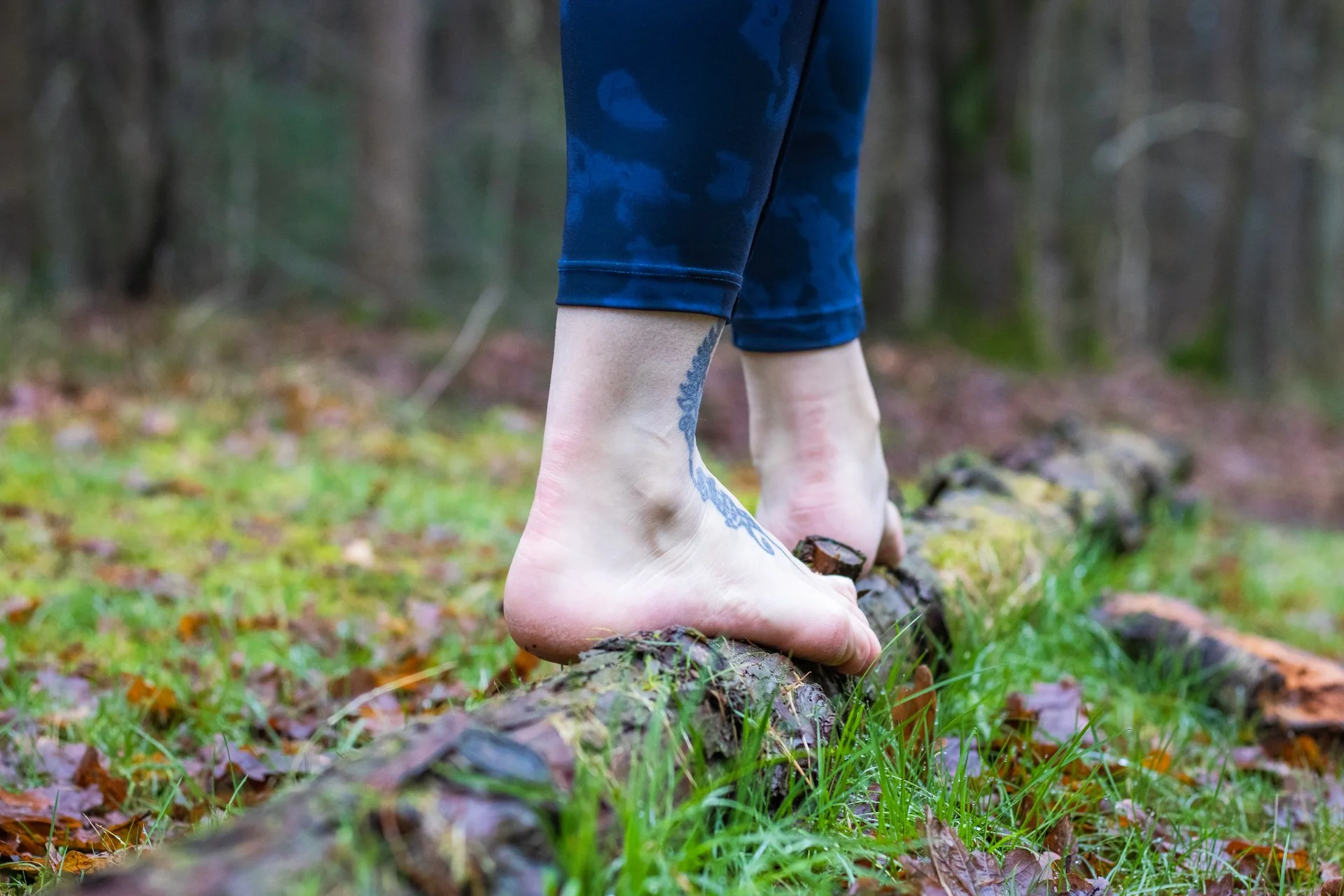 Person barefoot balancing on a fallen log in a forest, with tattooed legs and blue pants.