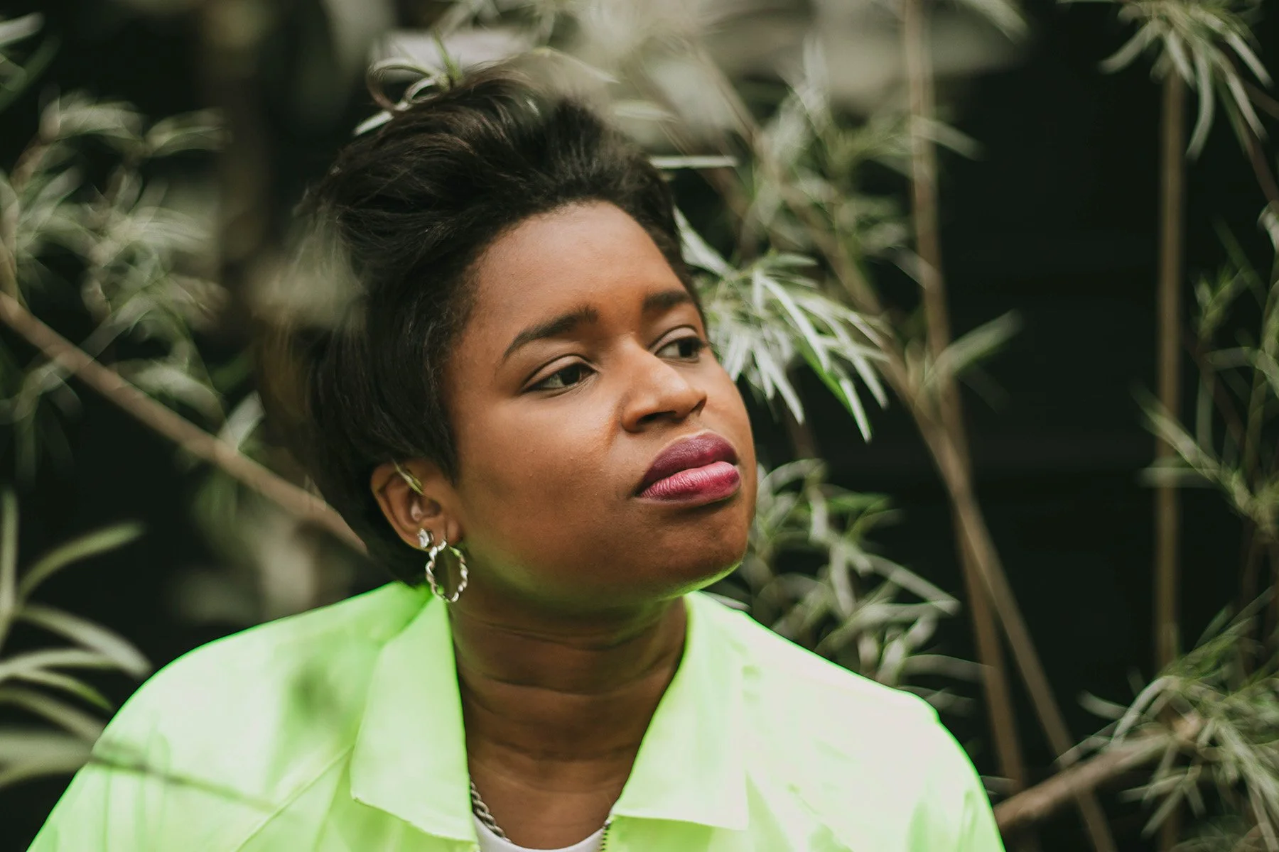A woman with short black hair, wearing a bright green jacket and hoop earrings, looking to her right with a neutral expression, with green leafy plants in the background.