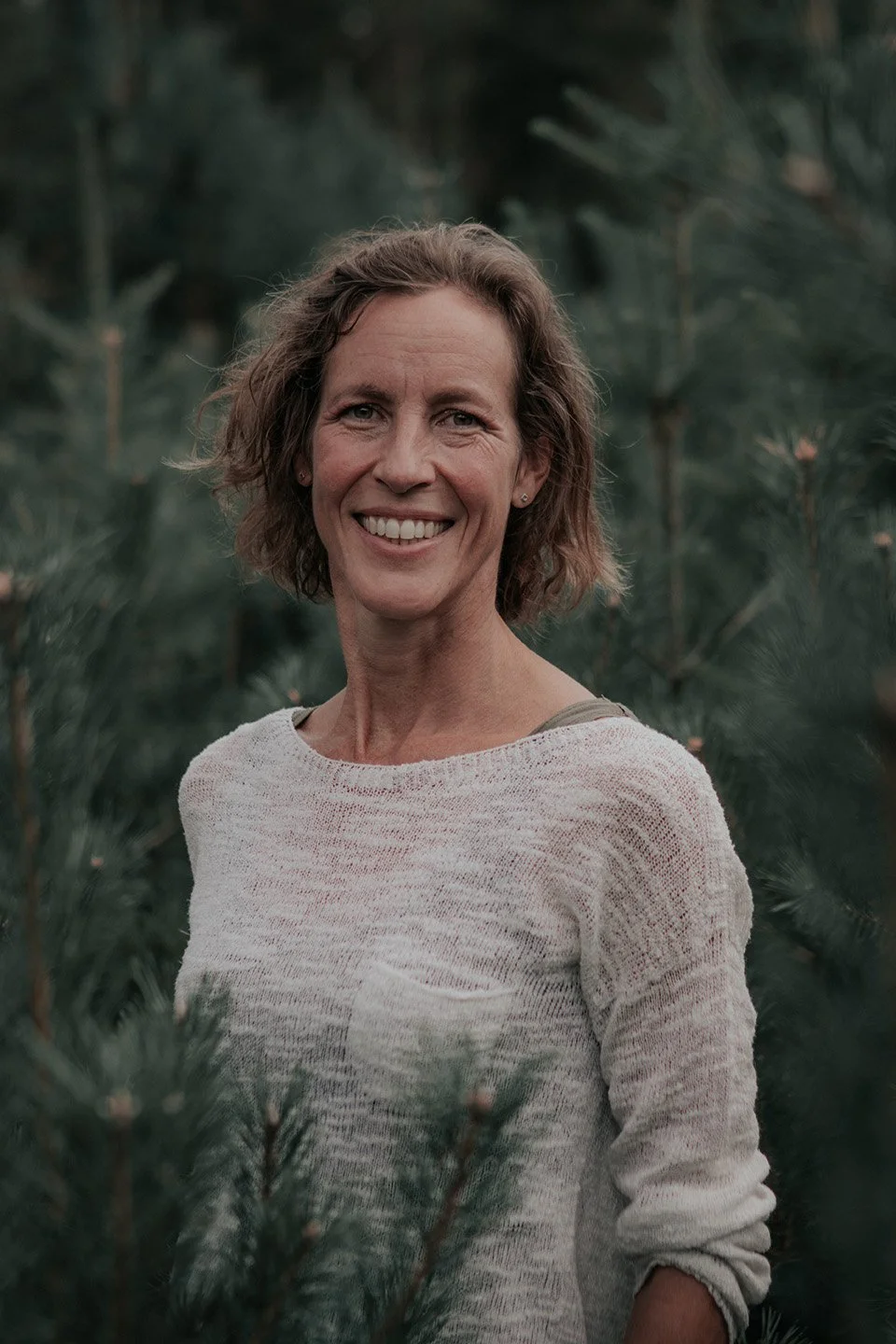 A smiling woman with short, wavy hair standing outdoors surrounded by green pine trees.