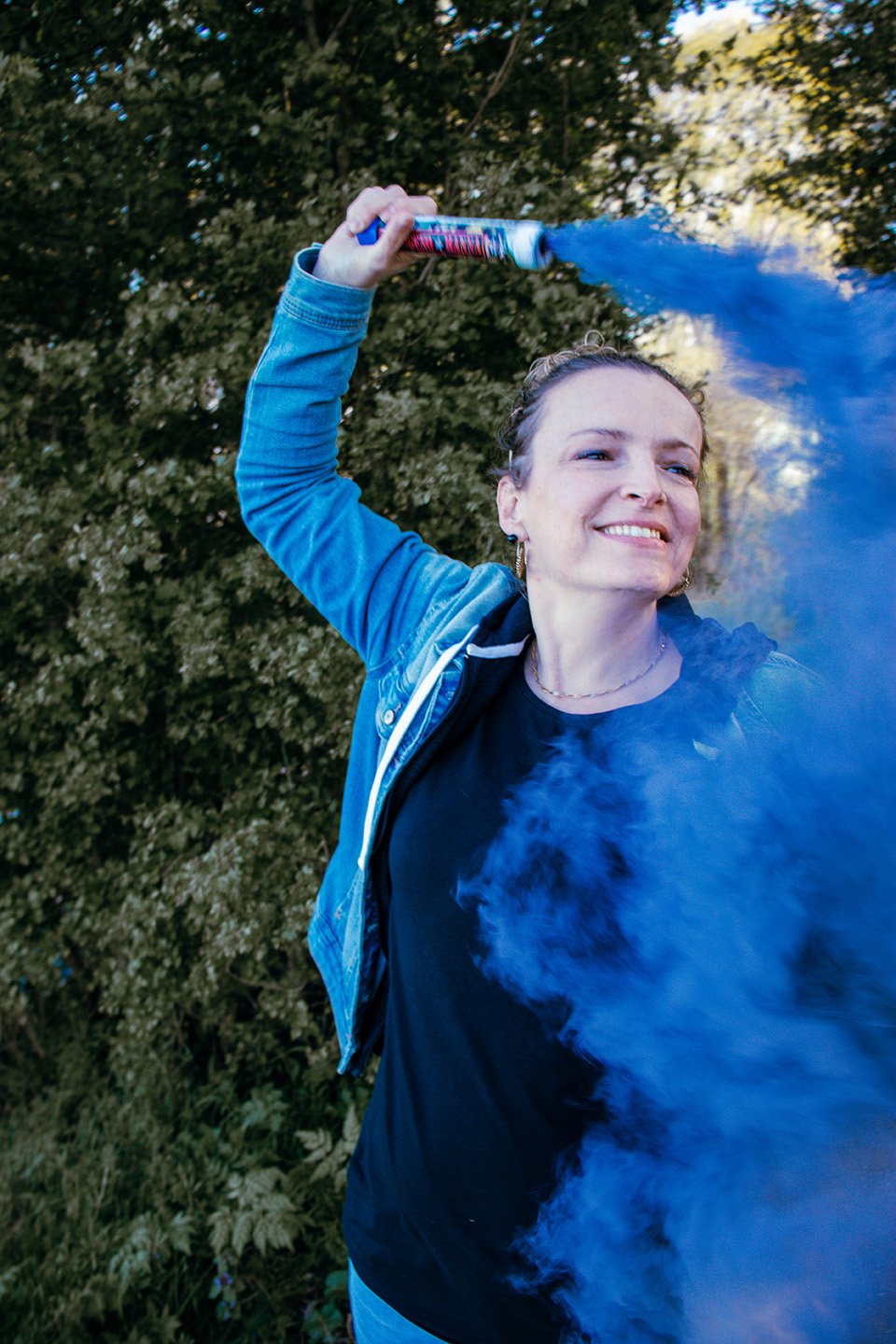 A woman smiling and holding a blue and red smoke bomb or pyrotechnic device, releasing blue smoke, in an outdoor setting with greenery and trees in the background.
