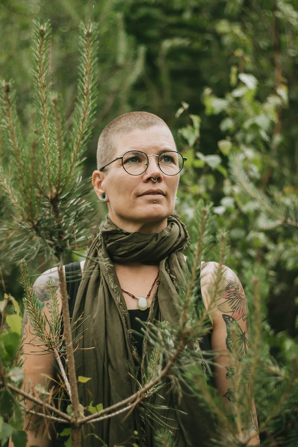 A person with glasses and tattoos, wearing a scarf, standing among green trees and plants in a forest.