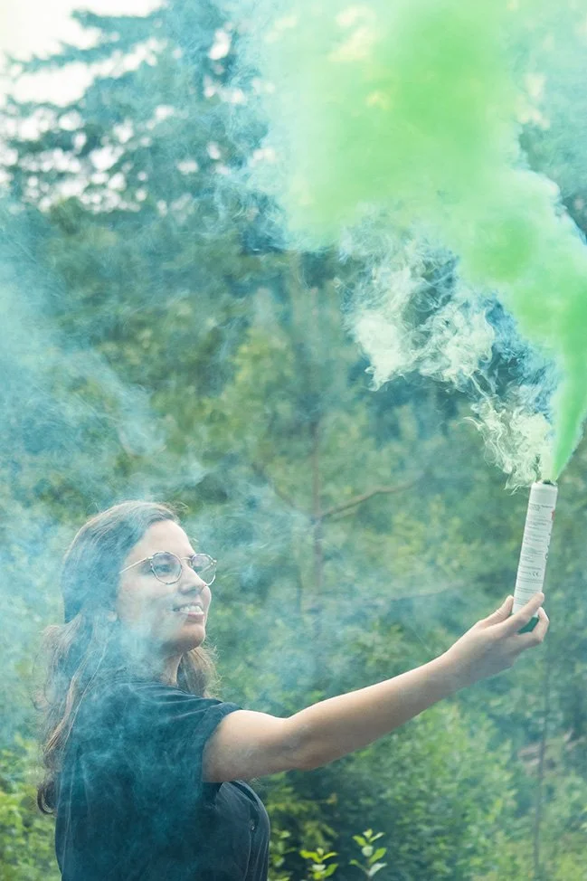 A woman holding a smoke grenade that releases green smoke outdoors, surrounded by trees and greenery.