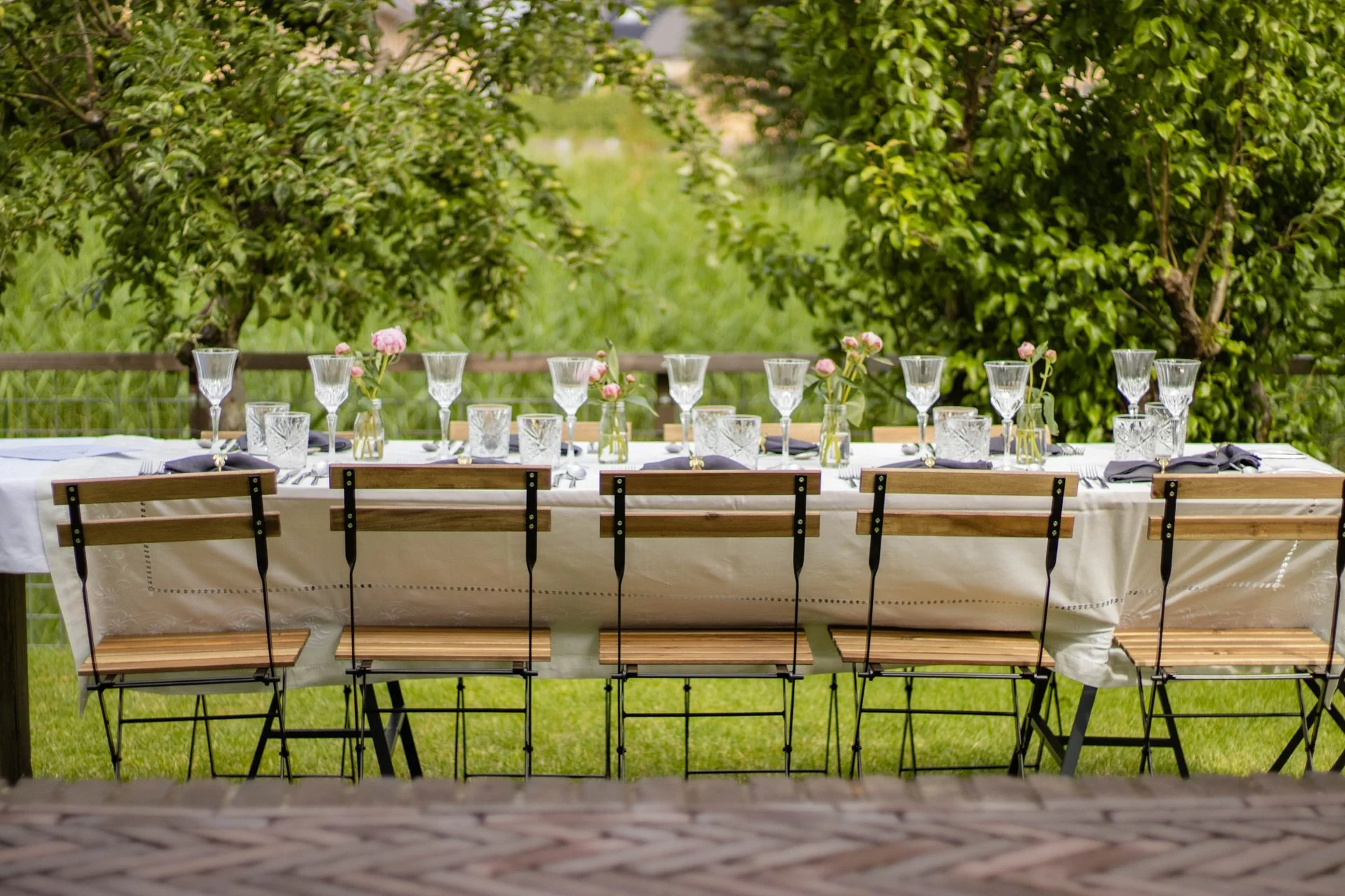 Set table for outdoor meal with glasses, candles, and pink flowers on a white tablecloth, surrounded by green trees and grass.