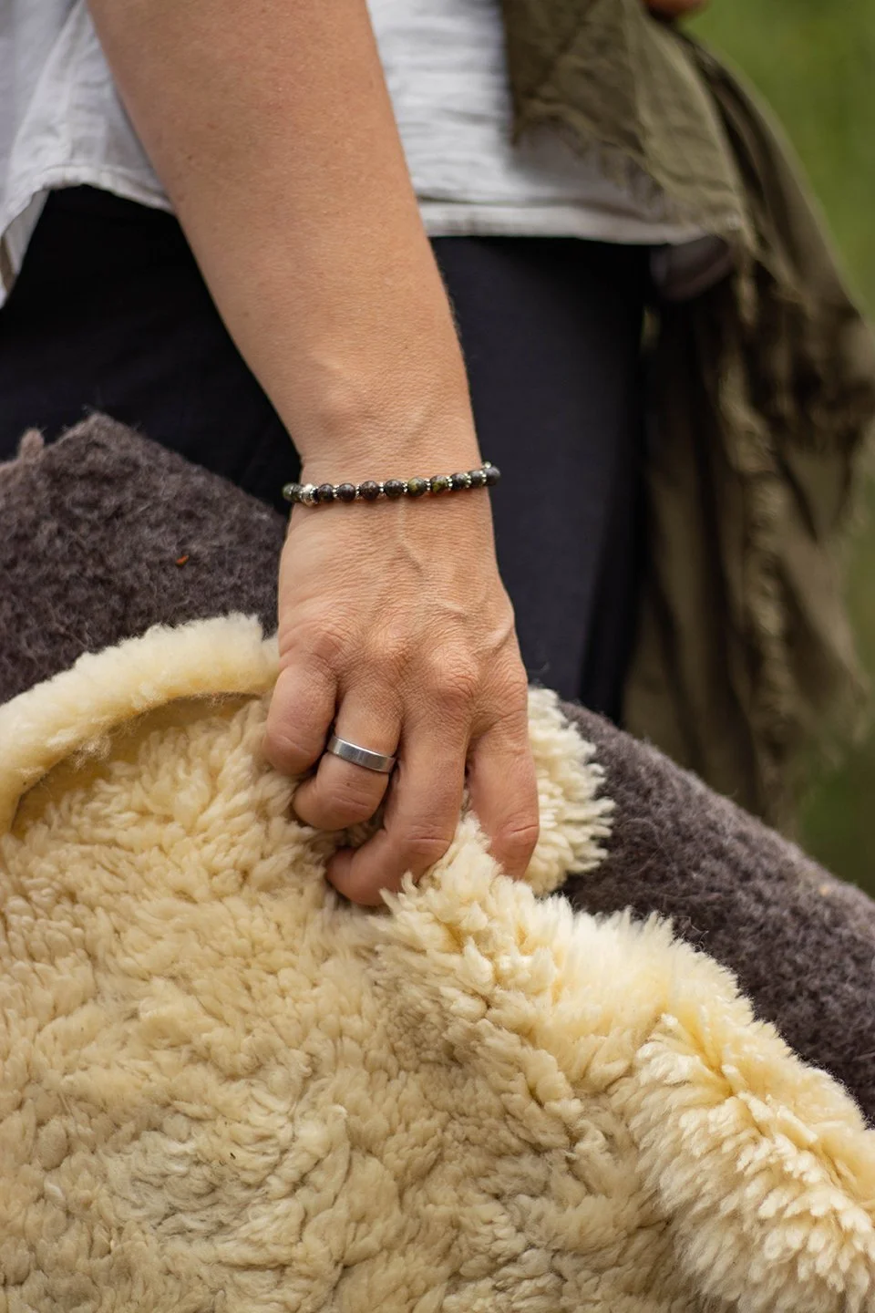 Close-up of a person's hand with a wedding band and beaded bracelet, resting on a fluffy cream-colored blanket.