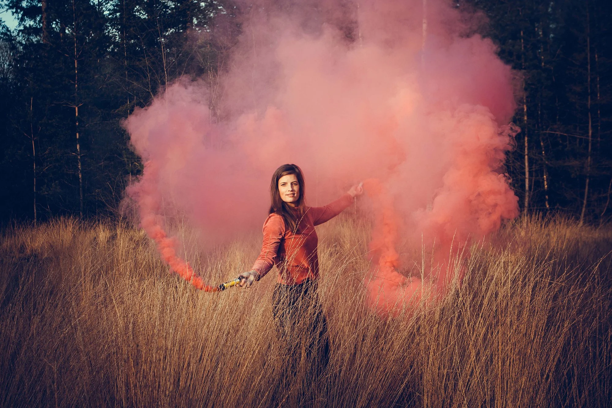 A woman standing in a field of tall grass, holding a pink smoke flare, with smoke billowing behind her during daytime.
