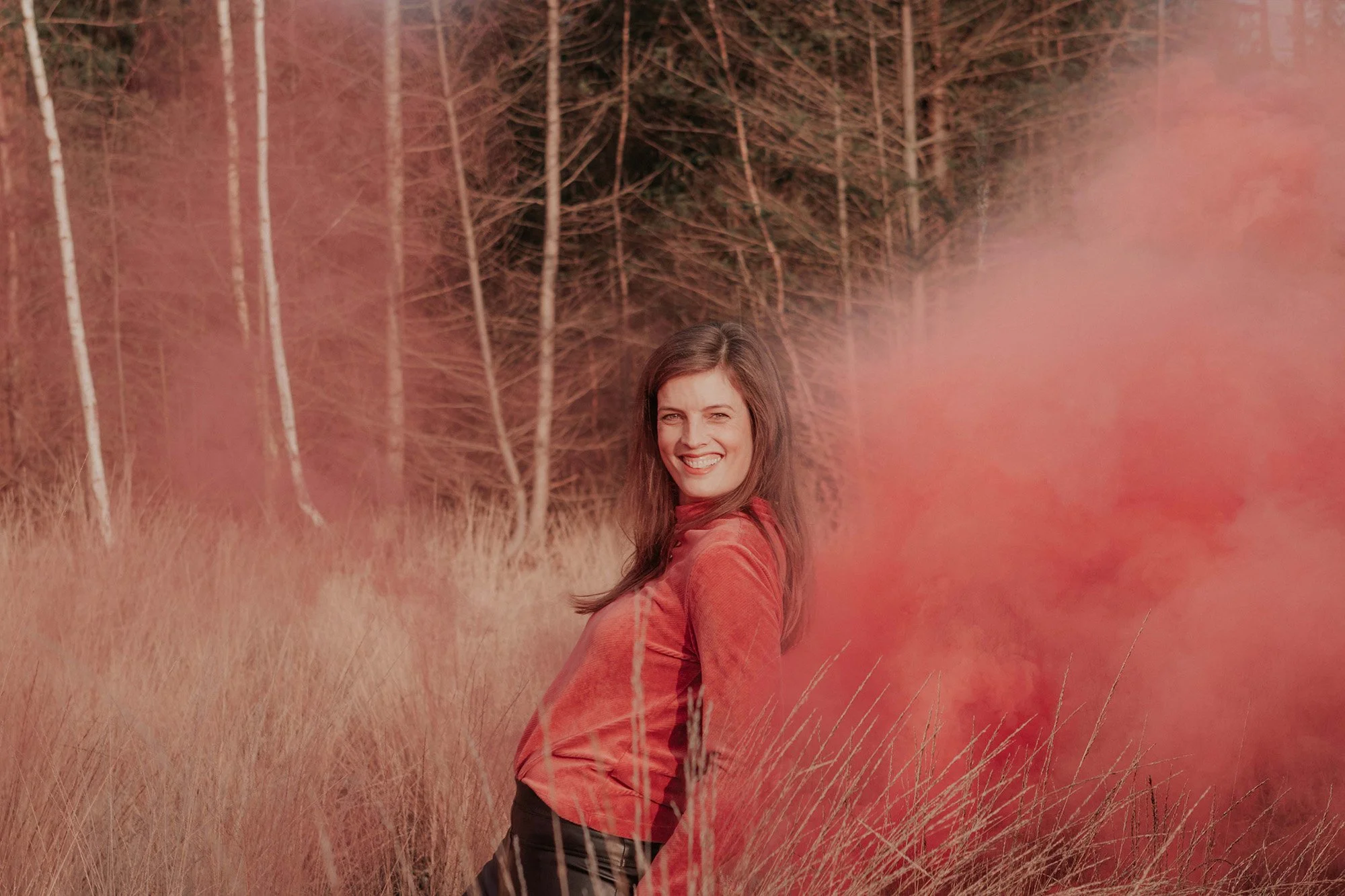 A woman smiling in a field of tall grass with pinkish-orange smoke or fog behind her, and a wooded forest area in the background.
