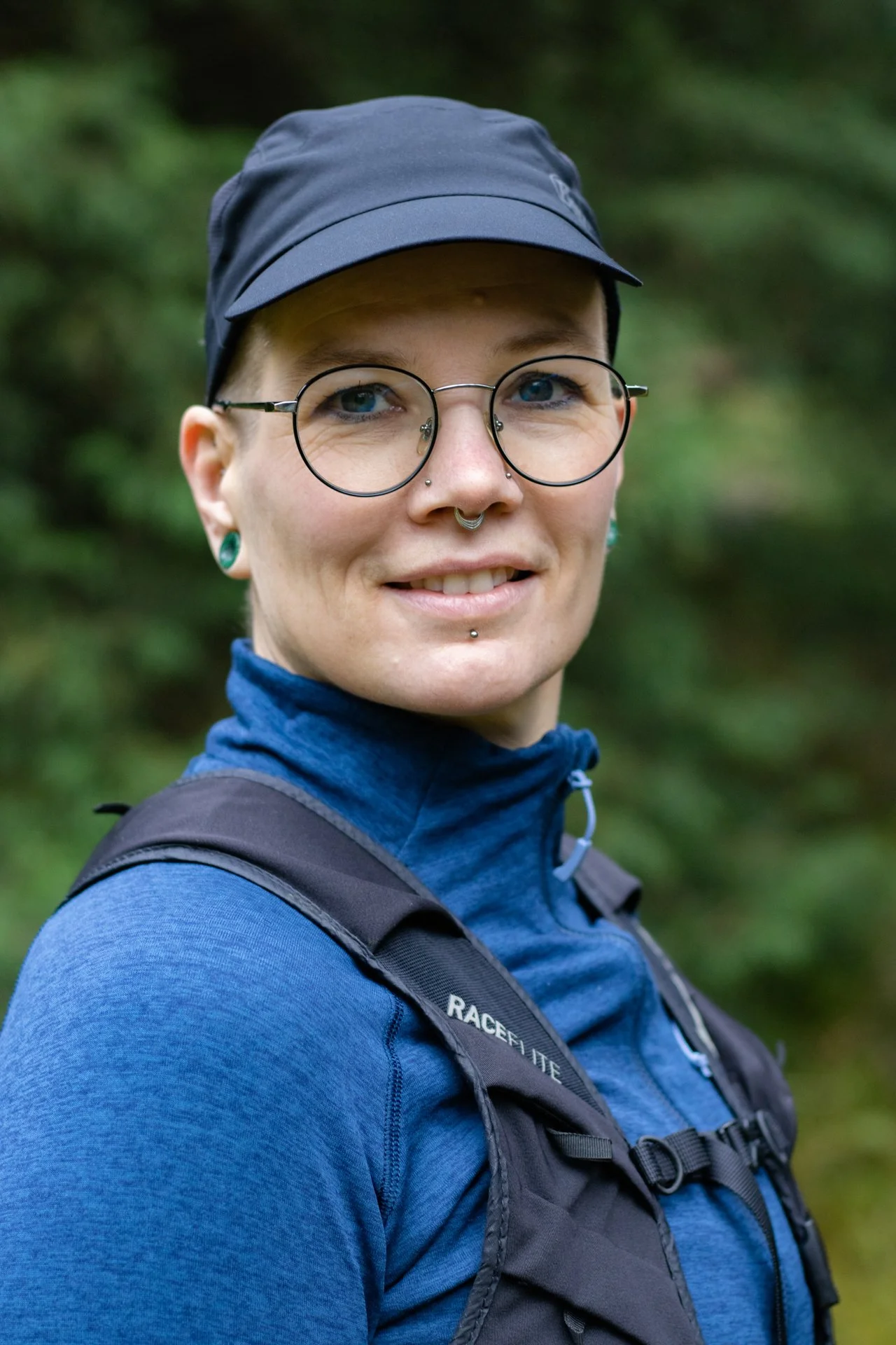 Portrait of a woman with glasses, piercings, and short hair, wearing a cap and a blue outdoor jacket, outdoors in a forested area.