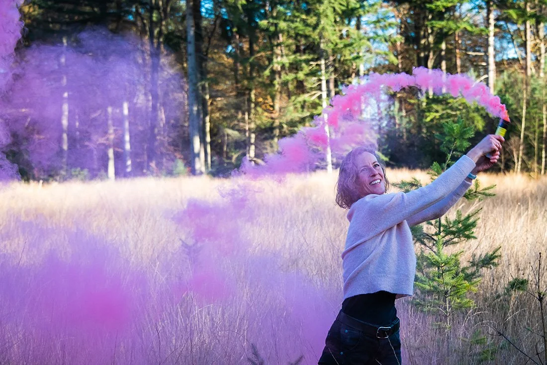 A woman in a gray sweater and black pants holding a pink smoke bomb in a forested field with trees in the background, creating a pink smoke ring.