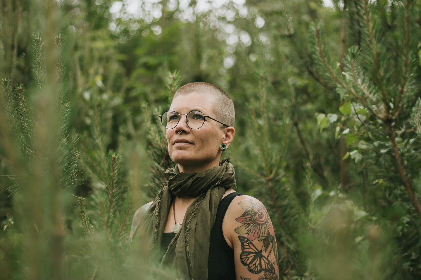 A woman with glasses and short hair stands among green trees with a tattoo of a butterfly and flowers on her arm, wearing a black top, a green scarf, and a necklace.