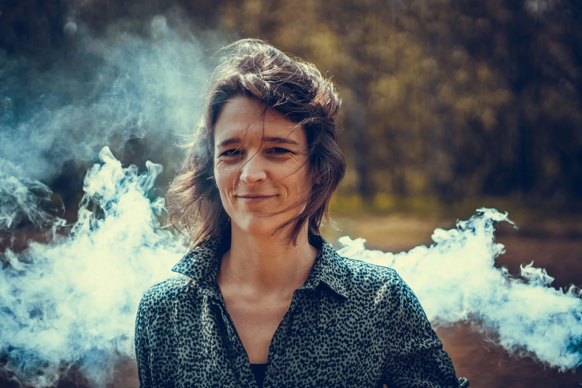 A woman with short, wavy hair standing outdoors with a smile, surrounded by smoke or mist, with trees or foliage in the background.