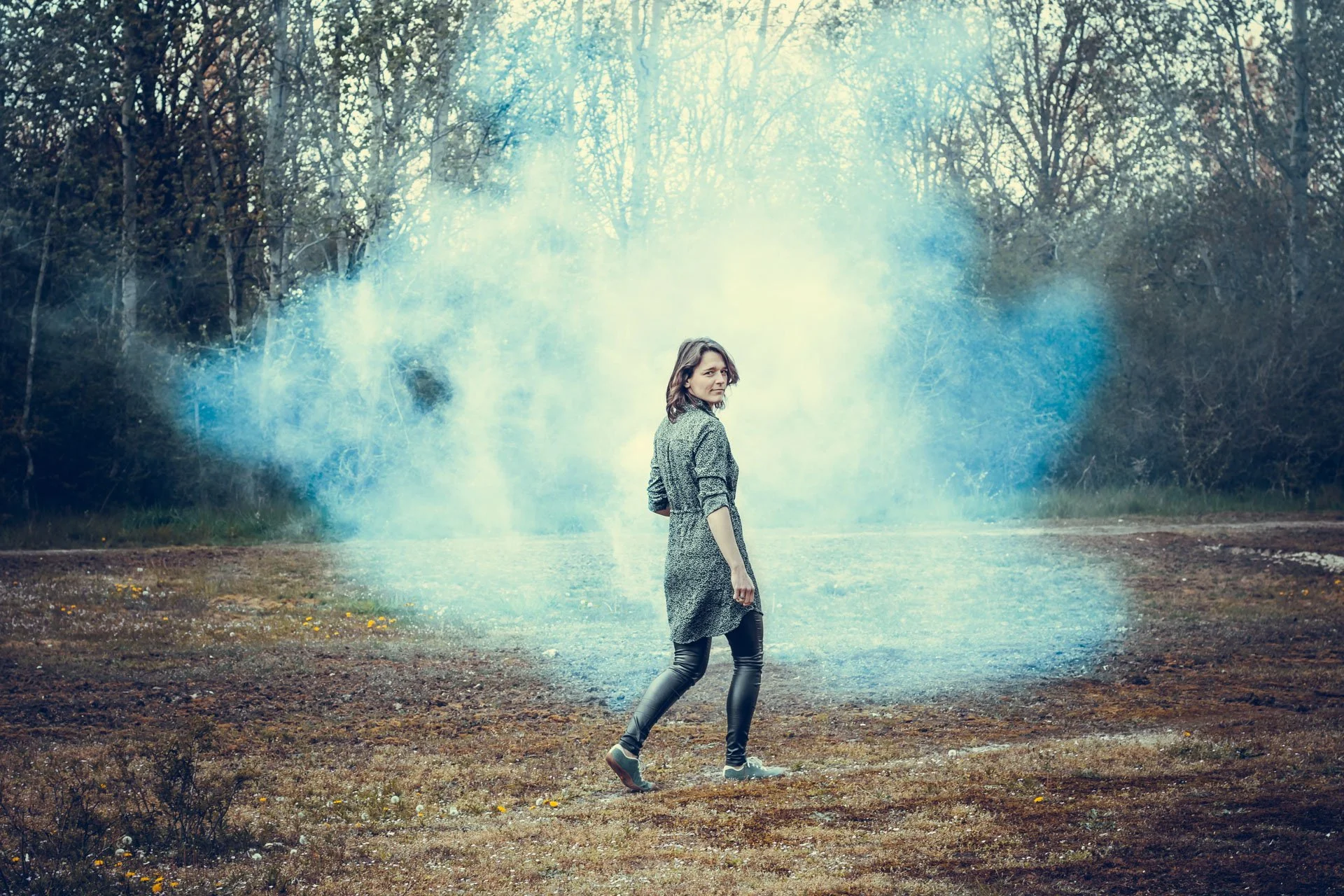 A woman walking outdoors on a dirt path surrounded by trees, with a cloud of blue smoke behind her.