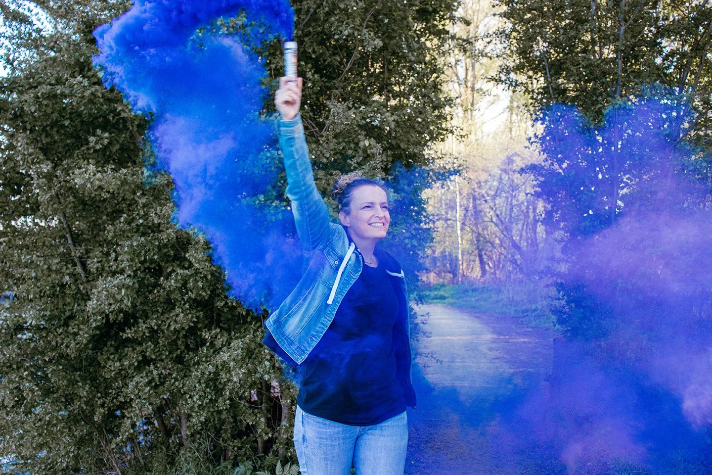 Woman smiling outdoors holding a blue smoke bomb in her right hand.