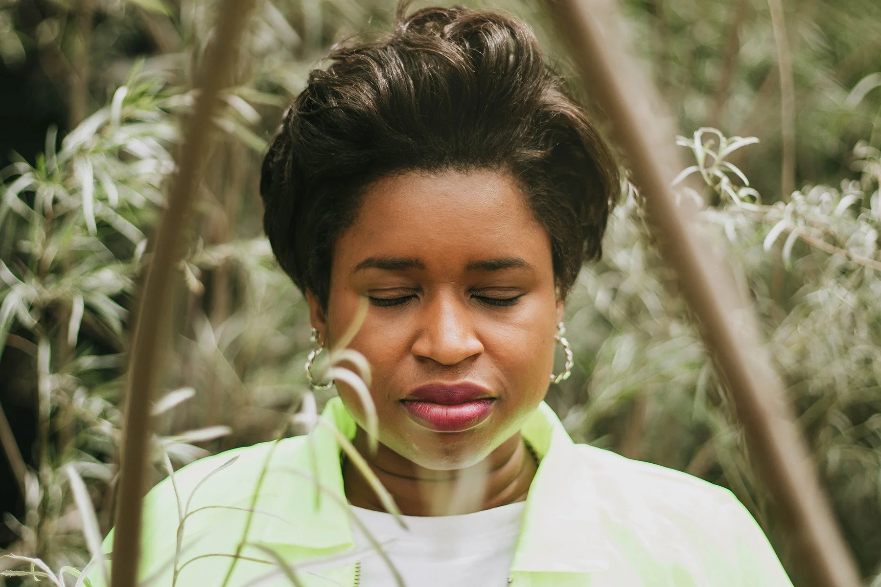 A woman with short dark hair and earrings standing among tall, thin plants with her eyes closed.