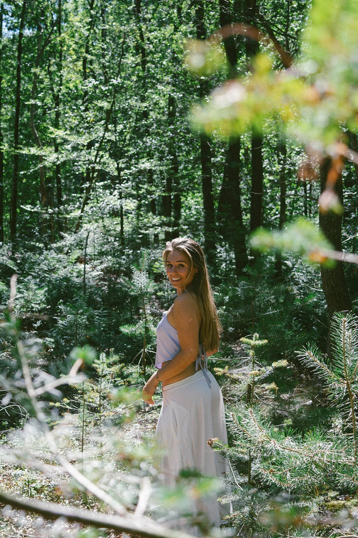 A woman with long hair smiling and standing in a dense forest with tall trees and lush green foliage.