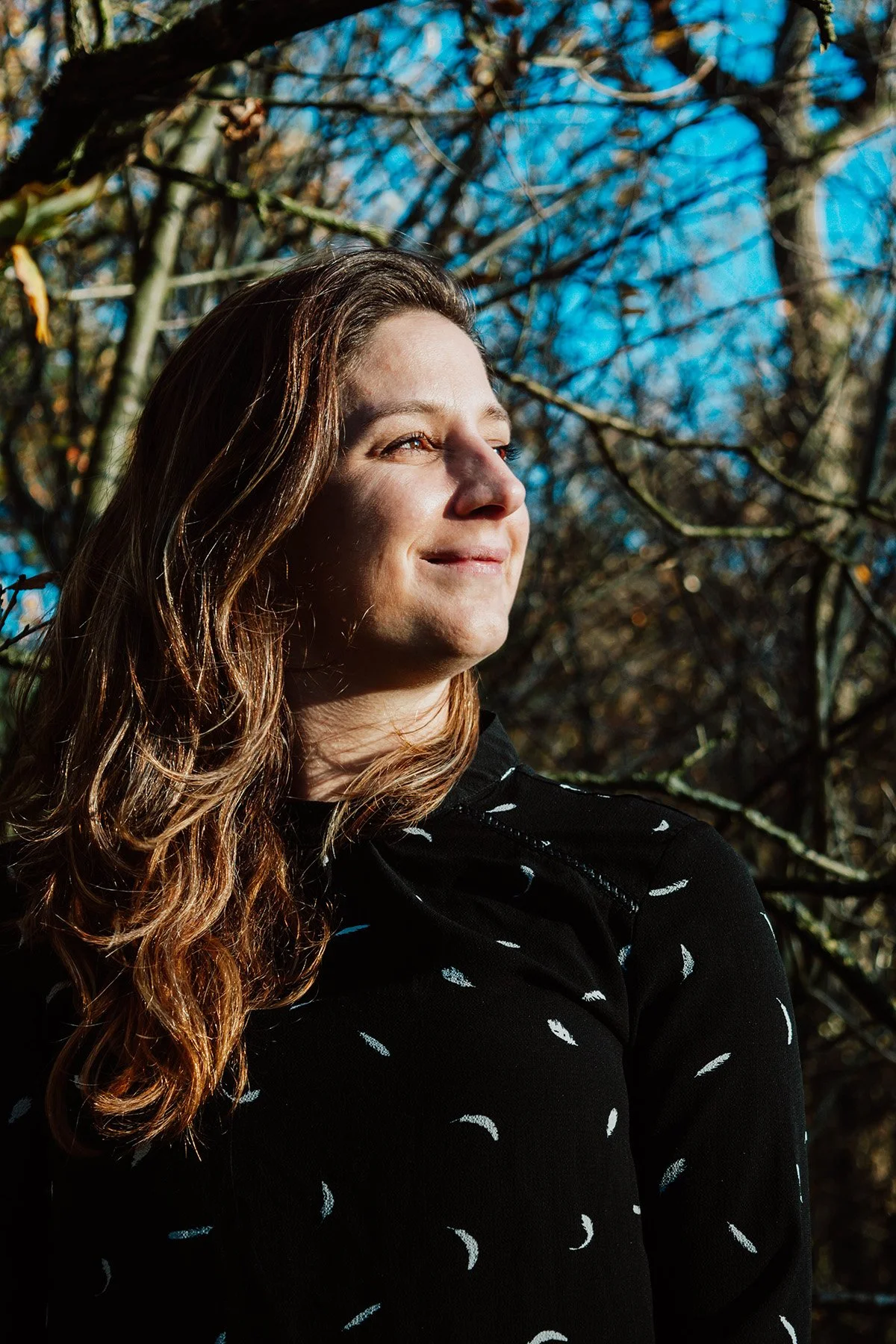 A woman with long, wavy brown hair smiling outdoors with trees and a blue sky in the background.