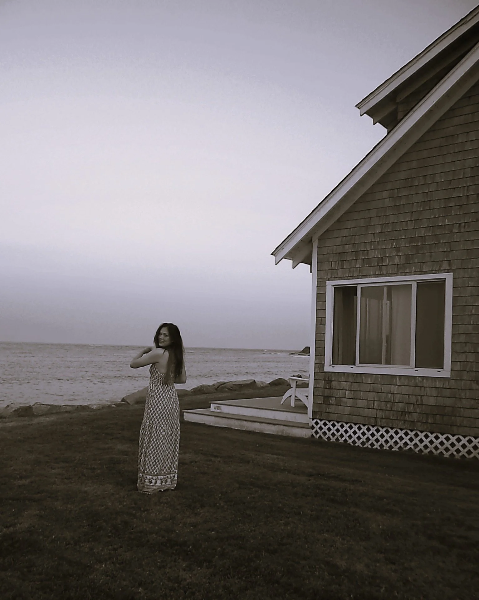 A woman in a long patterned dress standing outside near a house by the water, with rocks and a deck, during overcast weather.