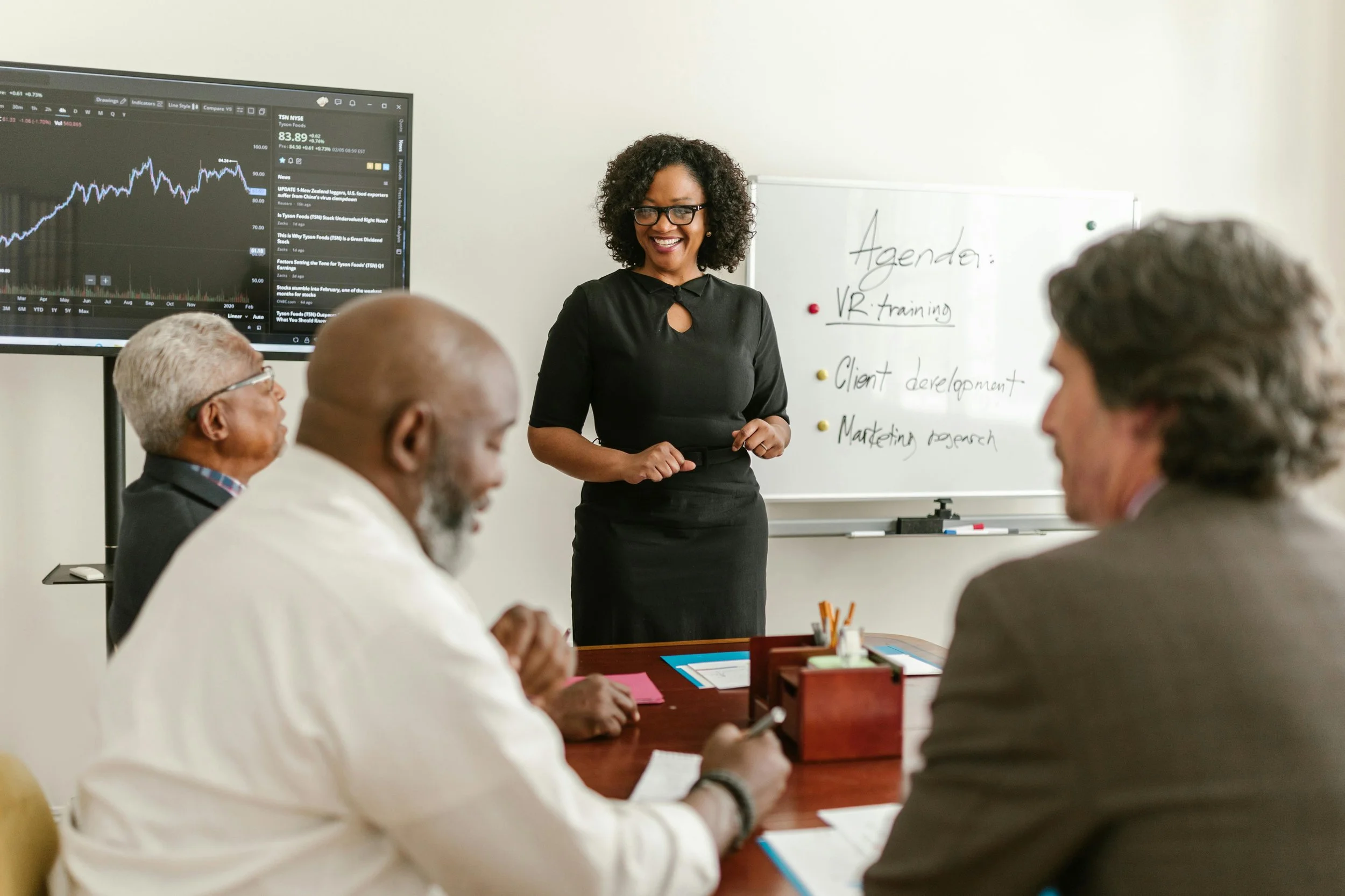 A woman presenter in a black dress giving a presentation to a diverse group of four people in a meeting room, with a whiteboard and a large screen displaying a stock market chart behind her.