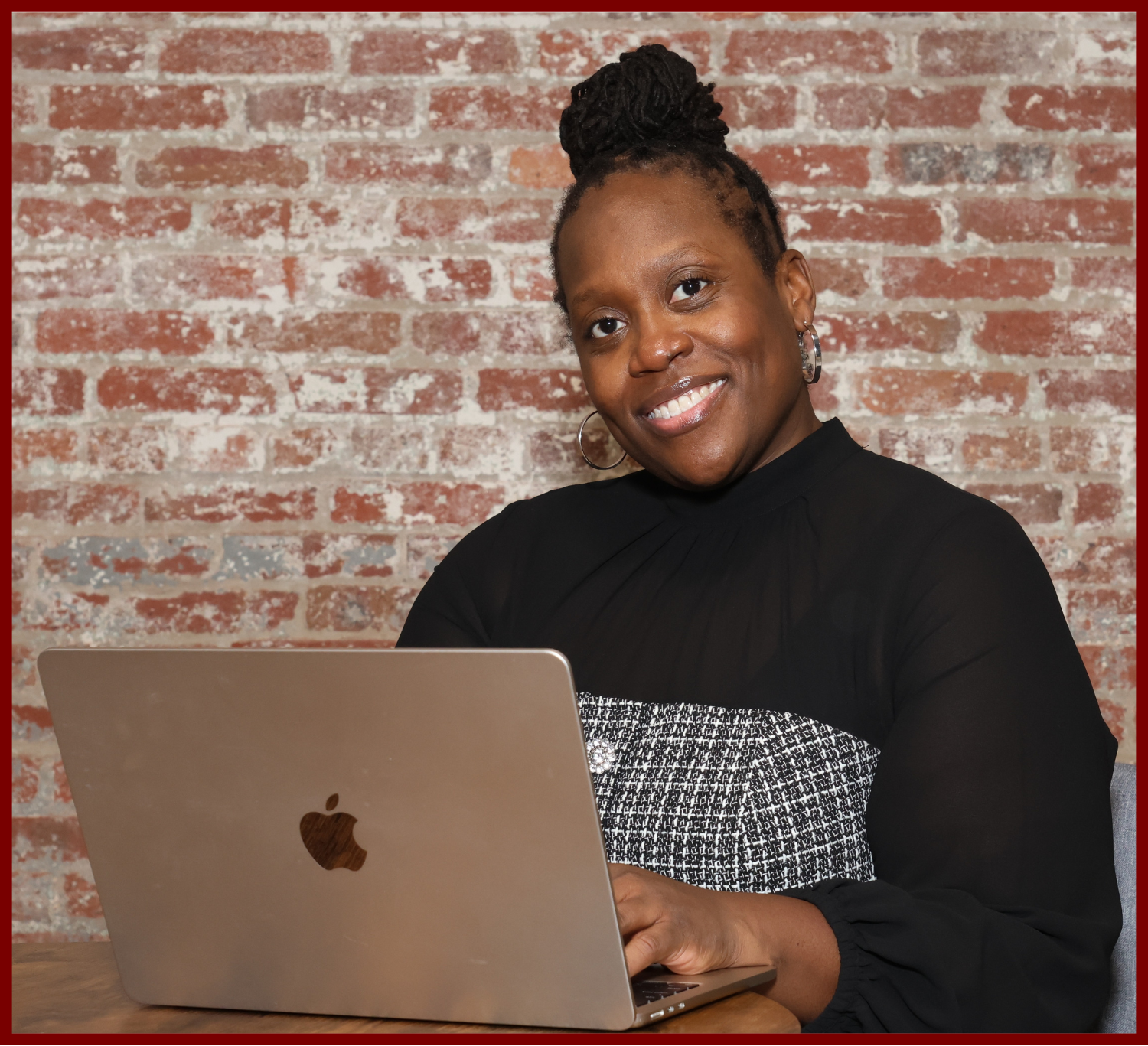 A woman with braided hair in a bun, smiling while working on a silver MacBook in front of a brick wall.