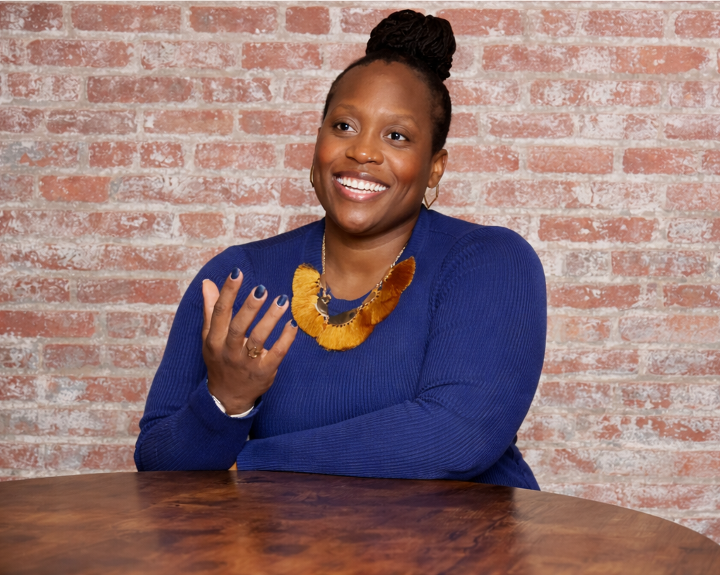 A woman smiling, wearing a blue sweater and a colorful necklace, sitting at a wooden table in front of a brick wall.