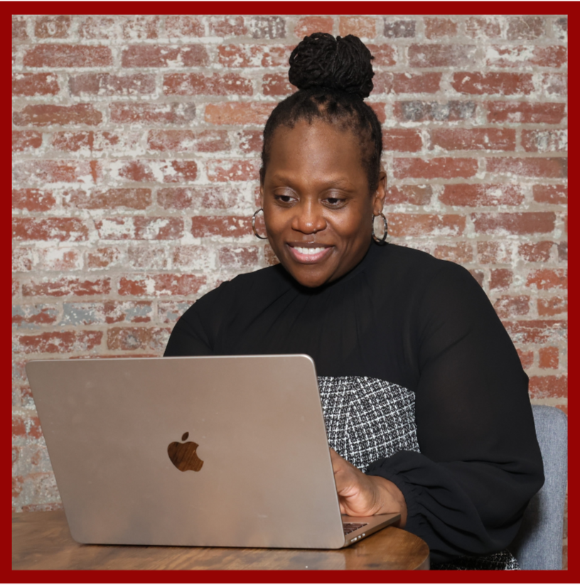 A woman with a high bun hairstyle using a silver MacBook laptop inside a room with an exposed brick wall.