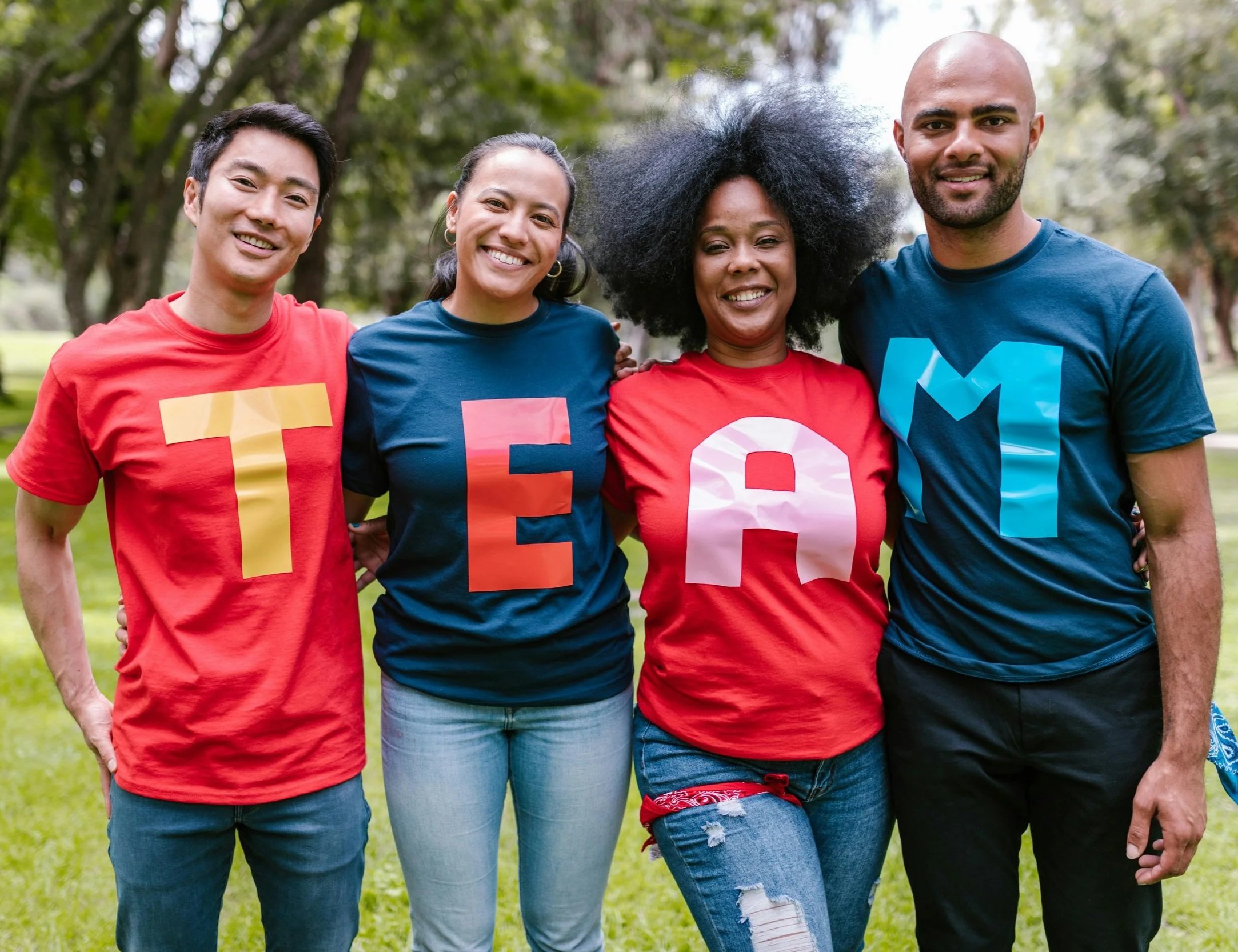 Four people standing outdoors, smiling and wearing colorful T-shirts with large letter prints spelling out 'TEAM'