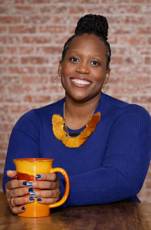 A woman with braided hair styled in a bun, smiling, wearing a blue sweater and a colorful yellow necklace, holding an orange mug, sitting in front of a brick wall.